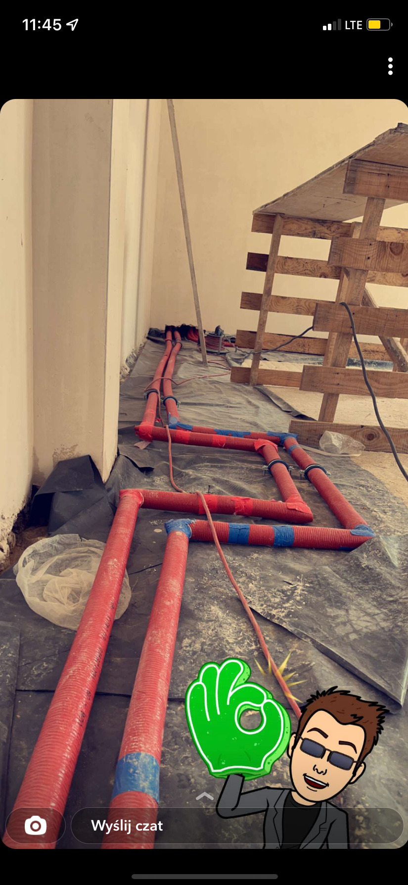 Czerwone rury ogrzewania podłogowego w izolacji, częściowo połączone i przymocowane taśmą, ułożone zygzakiem na czarnej folii, widoczny fragment drewnianej konstrukcji w surowym stanie i ściana...