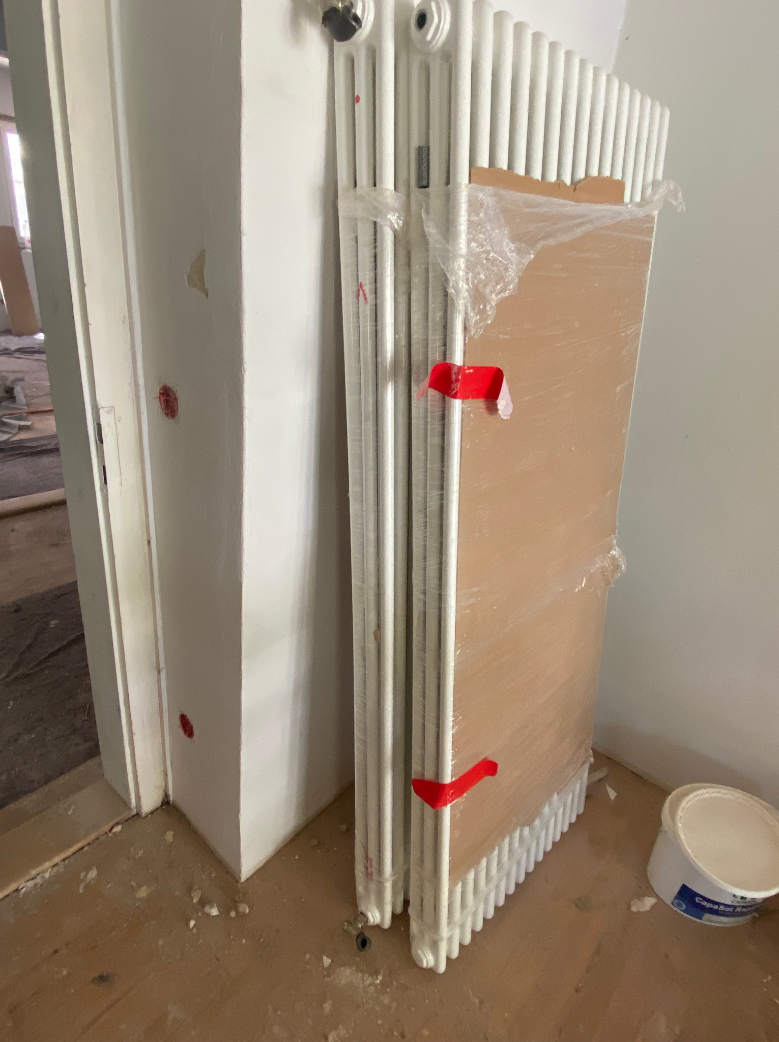 Białe, żeberkowe grzejniki owinięte folią ochronną, zabezpieczone czerwoną taśmą, oparte o ścianę obok futryny, z widocznym wiadrem z farbą na podłodze w trakcie remontu.