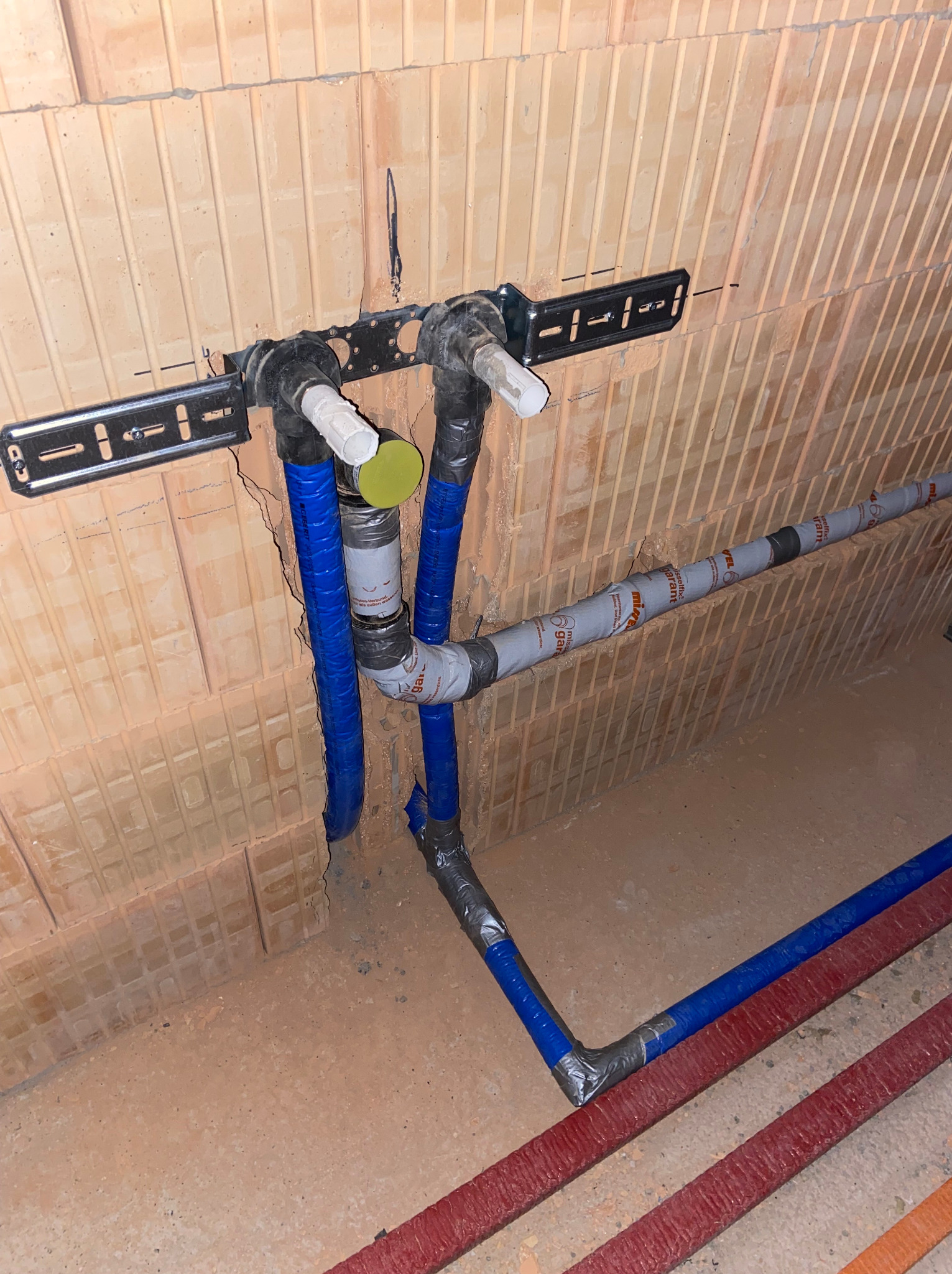 Instalacja wodna z rur PEX w izolacji, zamocowana do ściany z cegły dziurawki, z widocznymi elementami łączeniowymi i wspornikami metalowymi.