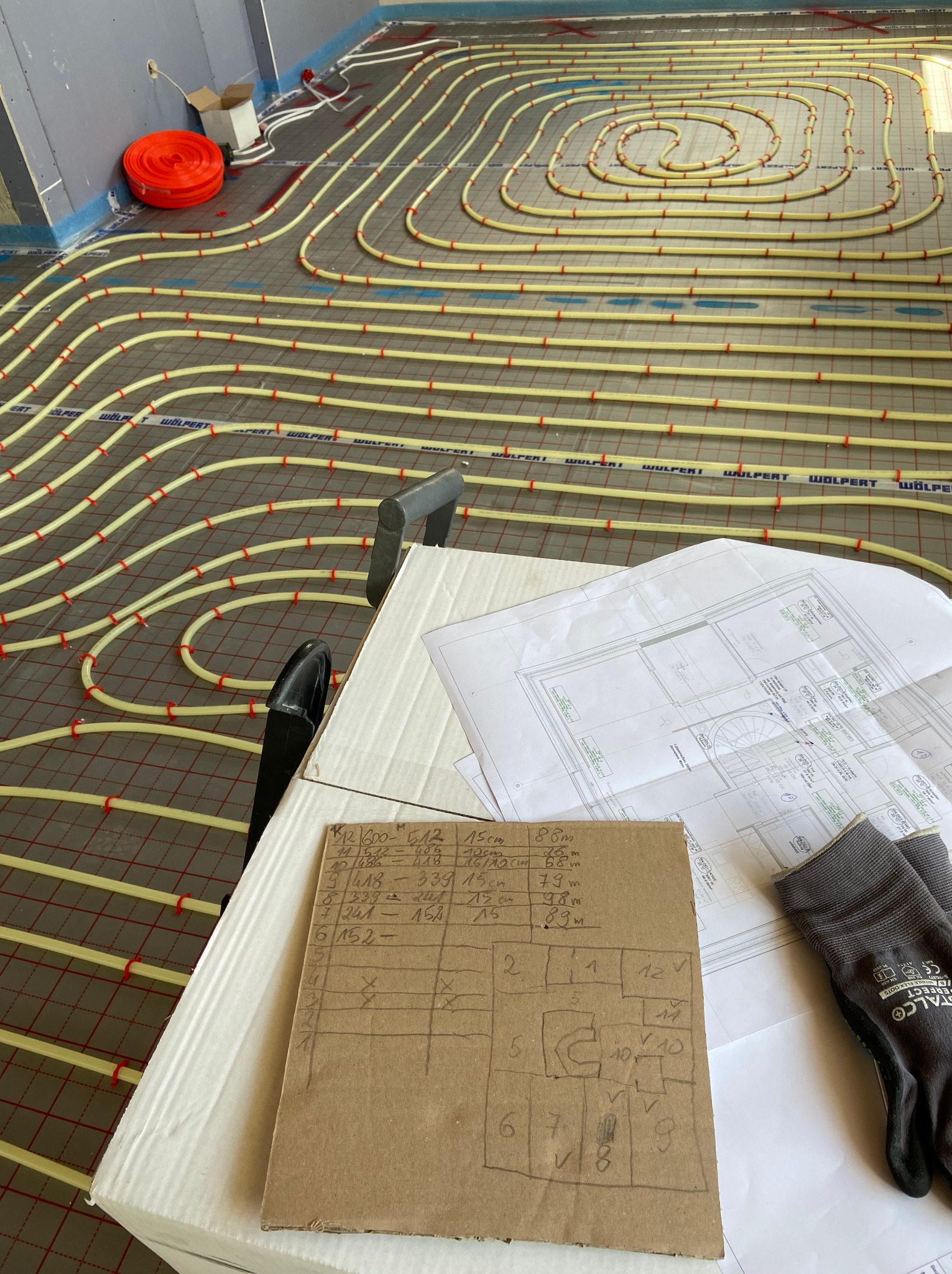 Układanie rur ogrzewania podłogowego w systemie pętli na siatce, widoczne plany instalacji, rękawice robocze i notatki na kartonie z rozrysowanym schematem pomieszczeń.