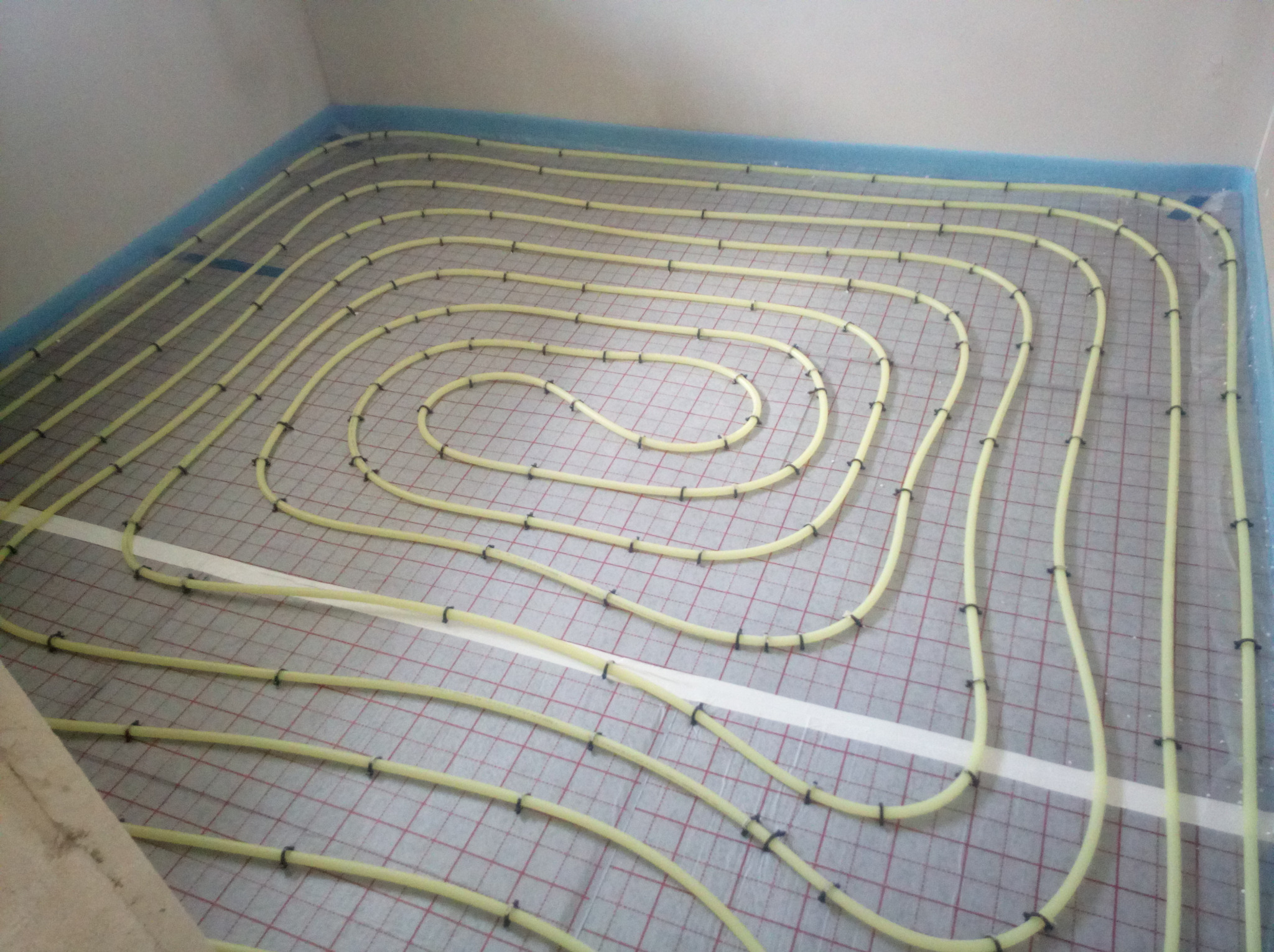 Żółte rury ogrzewania podłogowego ułożone spiralnie na folii z siatką, przymocowane czarnymi klipsami w pomieszczeniu z niebieską izolacją przy ścianach.