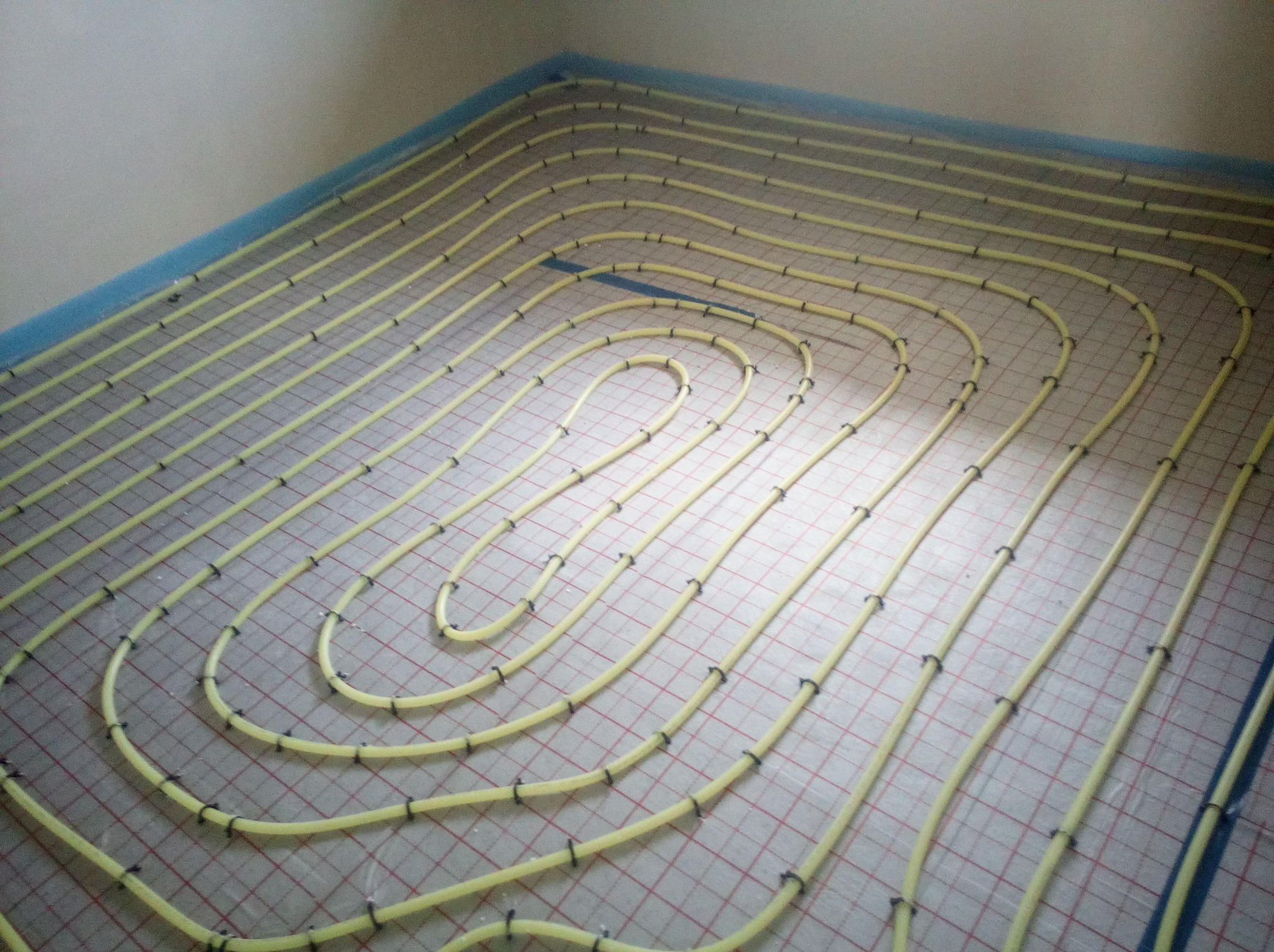 Żółte rury ogrzewania podłogowego ułożone w pętle na siatce montażowej, przymocowane czarnymi klipsami, widoczne przygotowania do wylewki.