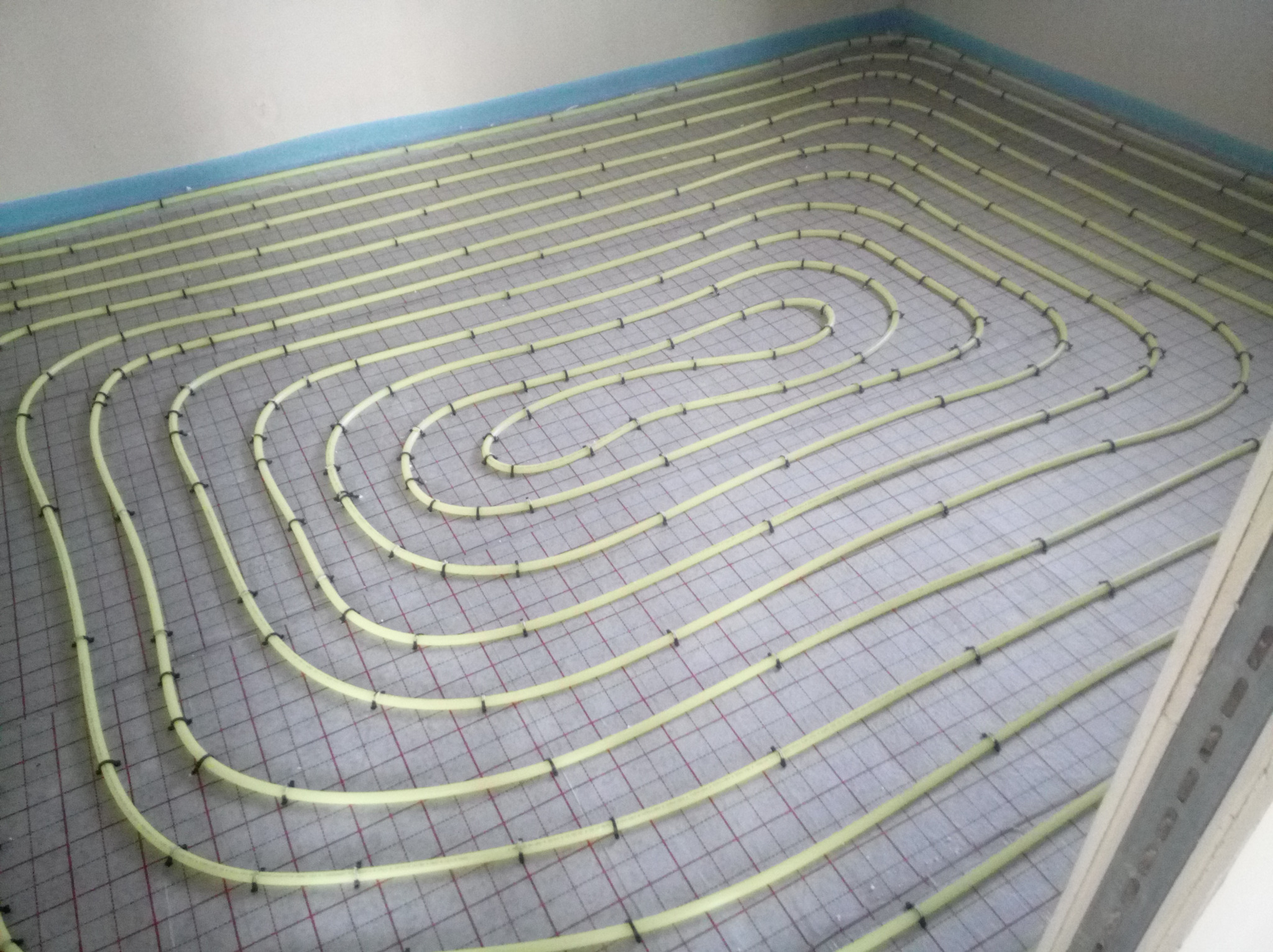 Żółte rury ułożone w pętle na siatce pod instalację ogrzewania podłogowego, zabezpieczone niebieską folią przy ścianie.