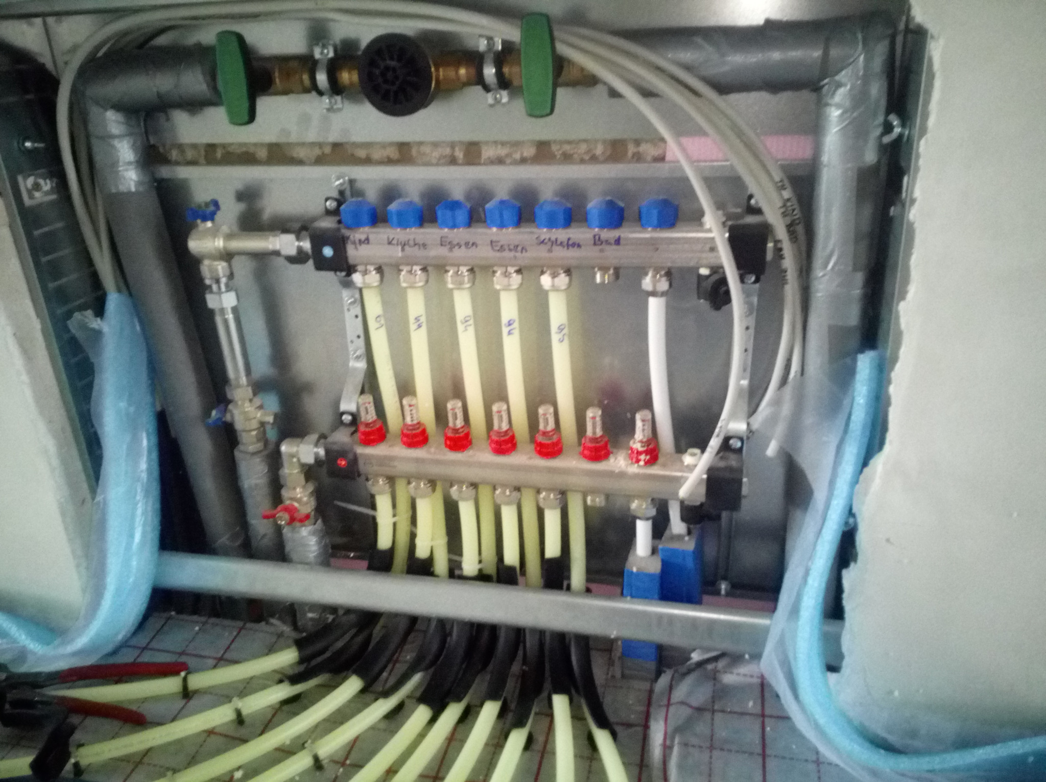 Rozdzielacz ogrzewania podłogowego z opisami pomieszczeń, widoczne rury doprowadzające i odprowadzające wodę, częściowo zamontowany system w trakcie instalacji.