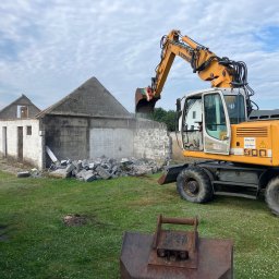 Świętokrzyskie Prace Ziemne Karol Kot - Żółta koparka Liebherr z łyżką kruszy resztki murowanego budynku w słoneczny dzień, widoczne gruzy i trawa.