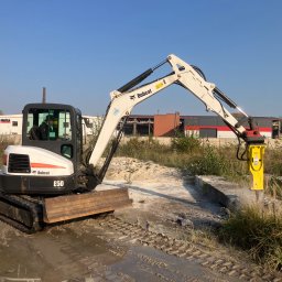 Świętokrzyskie Prace Ziemne Karol Kot - Mini koparka Bobcat E50 z hydraulicznym młotem pneumatycznym rozbija betonowy element na placu budowy, widoczne ślady gąsienic w błocie i budynki w tle.