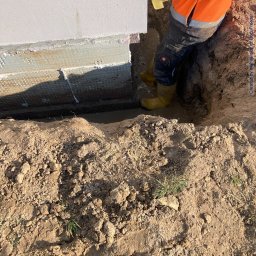 Świętokrzyskie Prace Ziemne Karol Kot - Pracownik w żółtych gumowcach stoi w wykopie przy ścianie budynku, trzymając żółty wąż; widoczna ziemia i fragment ocieplenia fundamentów.