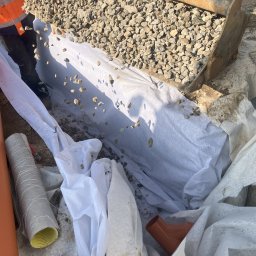 Świętokrzyskie Prace Ziemne Karol Kot - Wykop wyłożony białą geowłókniną, częściowo zasypany żwirem. Widoczne rury drenarskie, pomarańczowe złączki i fragment koparki.