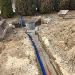 Wykop w ziemi z ułożonymi rurami instalacyjnymi w kolorze niebieskim i pomarańczowym, widoczny betonowy zbiornik z pokrywą, mini koparka i taczka w tle, teren przygotowany pod instalacje.