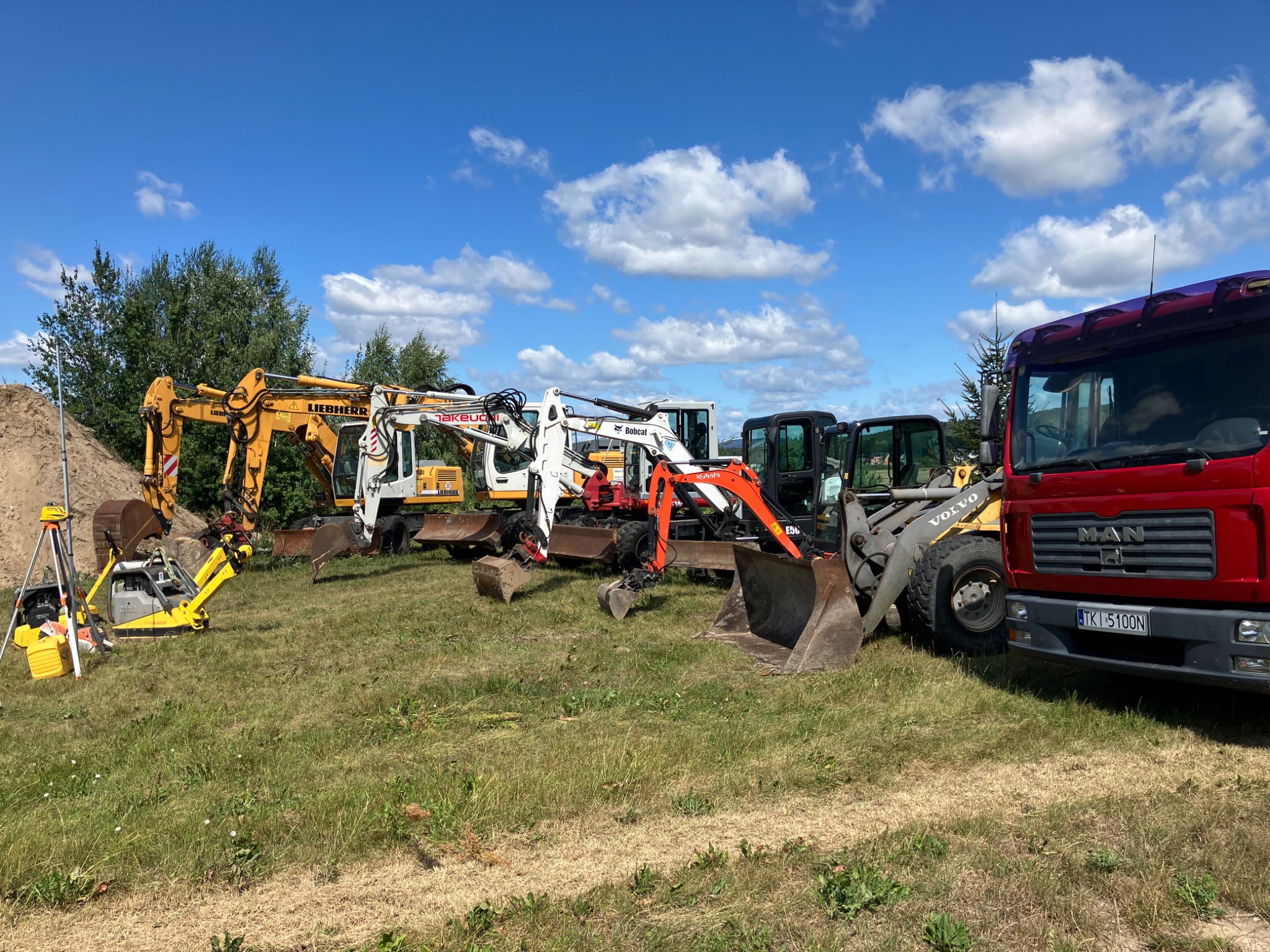 Sprzęt budowlany: żółta ubijarka gruntu, żółta niwelatorka na statywie, koparki Liebherr, Takeuchi, Bobcat i Kubota oraz ładowarka Volvo i czerwona ciężarówka MAN, ustawione na trawiastym terenie...