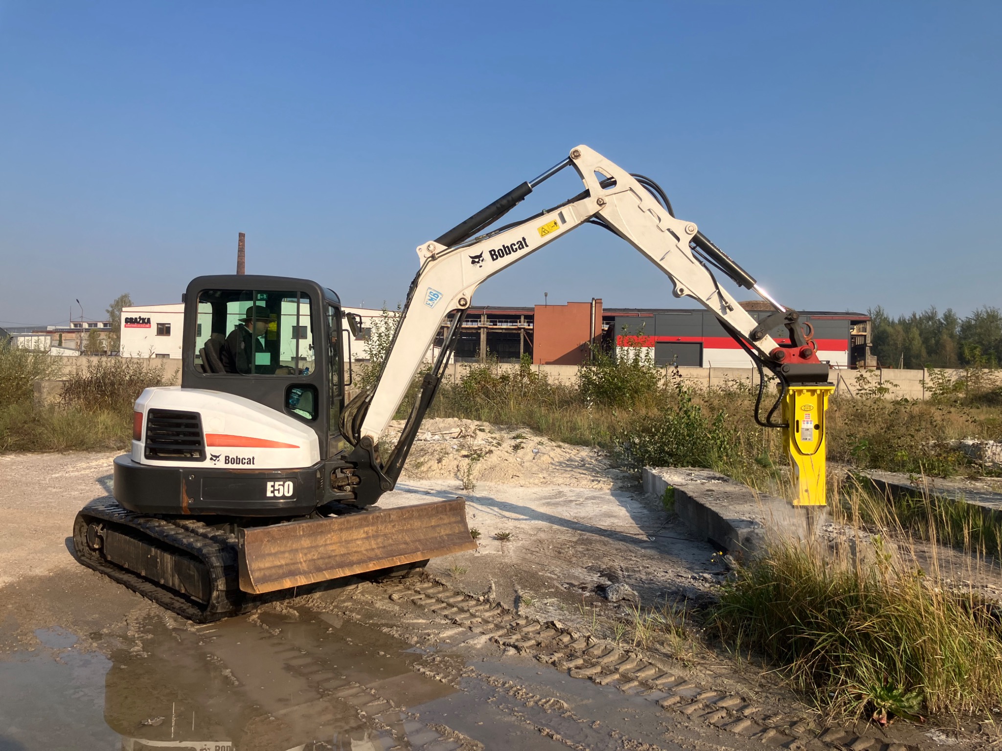 Mini koparka Bobcat E50 z hydraulicznym młotem pneumatycznym rozbija betonowy element na placu budowy, widoczne ślady gąsienic w błocie i budynki w tle.