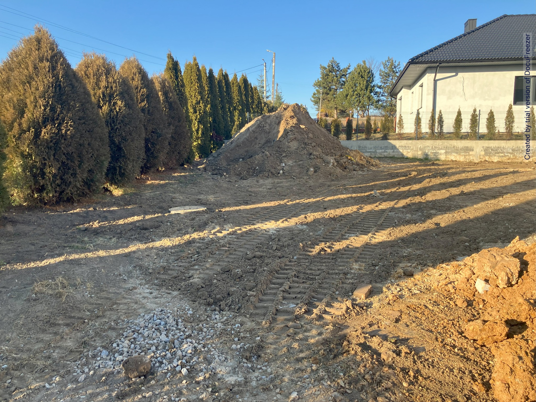Kupa ziemi na placu budowy, widoczne ślady opon maszyny budowlanej, w tle rząd tuj i dom jednorodzinny.