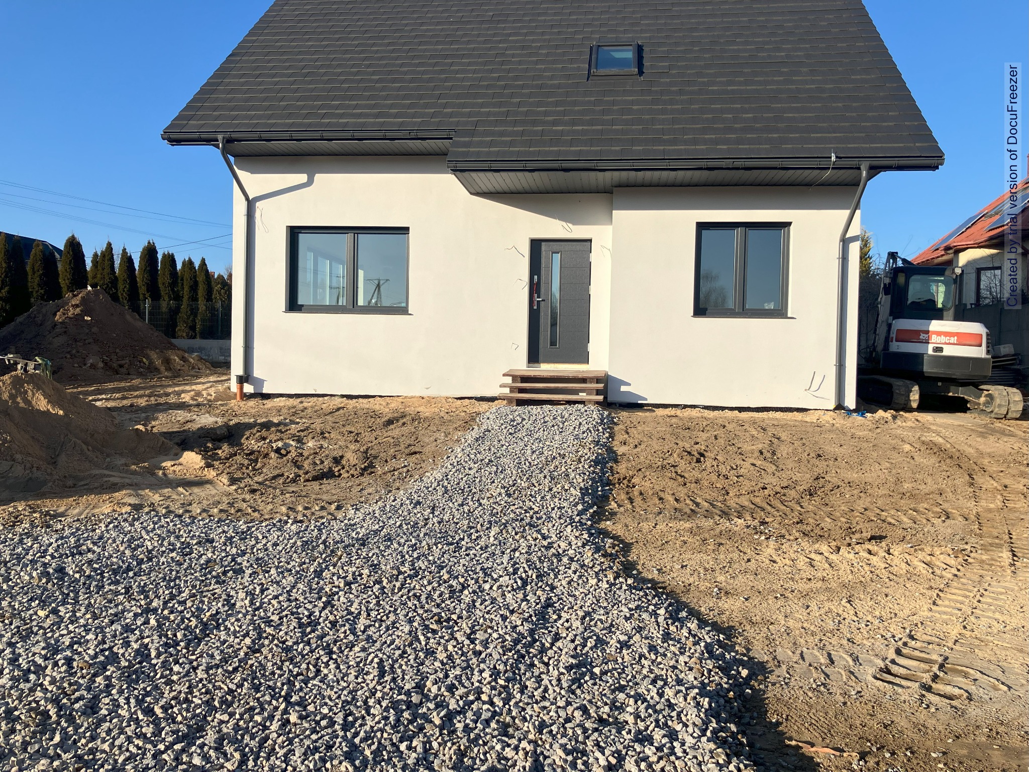 Nowo wybudowany dom jednorodzinny z szarą kostką prowadzącą do wejścia, teren wokół domu w trakcie niwelacji, widoczna mini koparka Bobcat w tle.