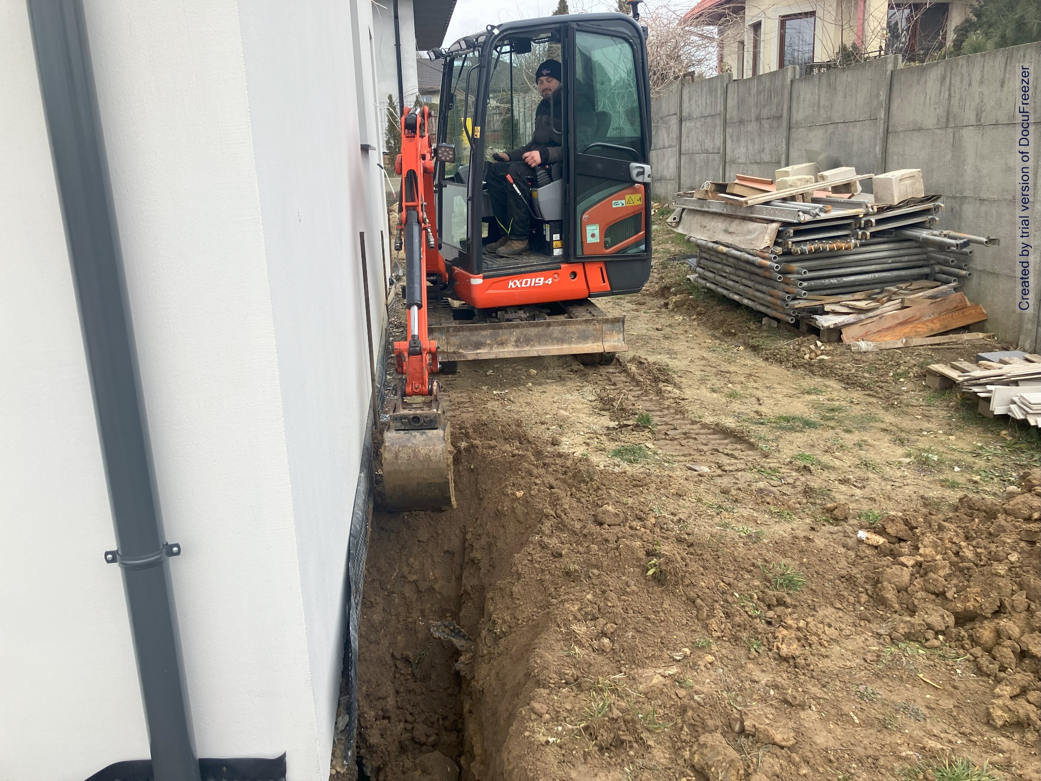 Pomarańczowa mini koparka Kubota KX019-4 z operatorem w czapce, wykonująca wykop wzdłuż białej ściany budynku, w tle stos rusztowań i betonowe ogrodzenie.