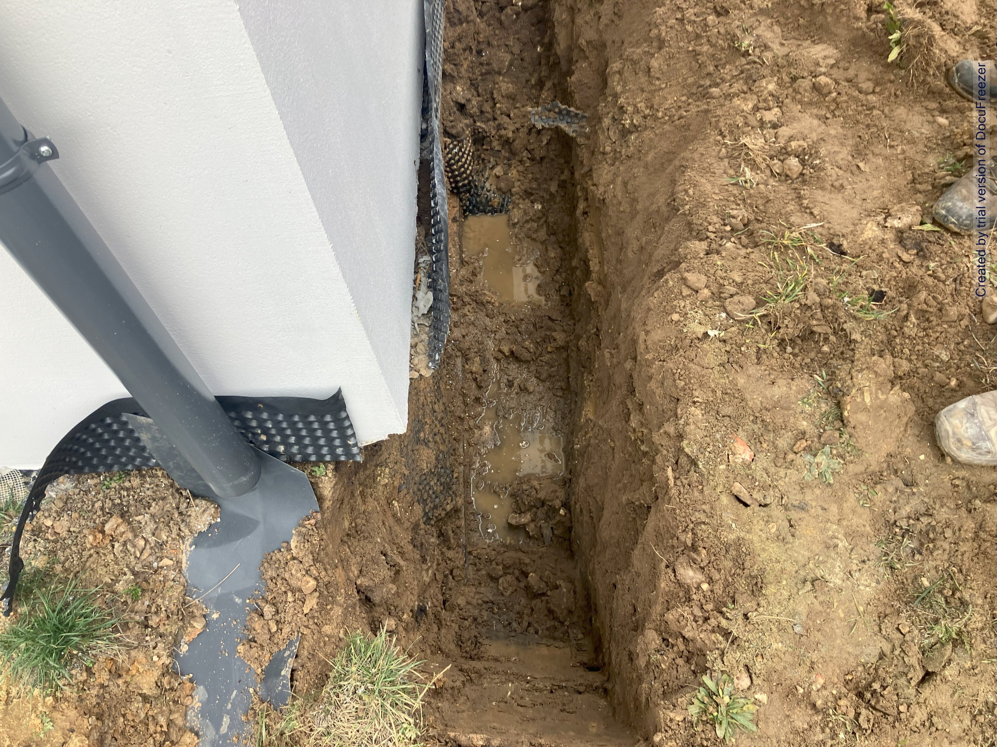 Wykopany wąski rów wzdłuż białej ściany budynku z widoczną rurą spustową, folią kubełkową i stojącą wodą na dnie wykopu.