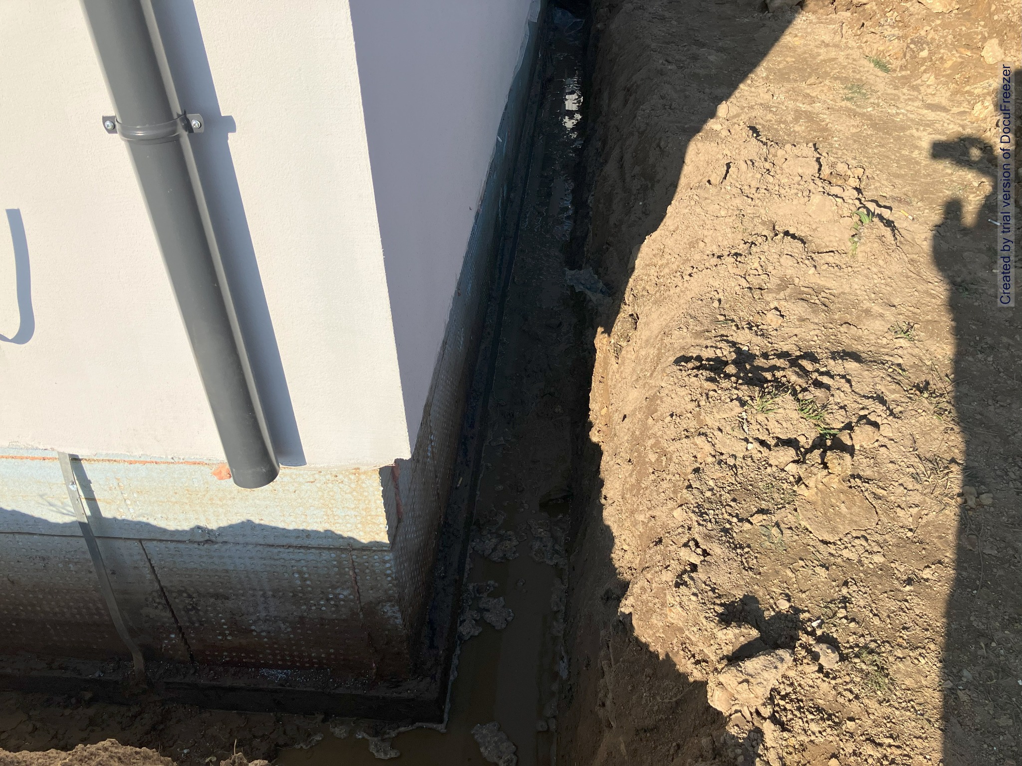 Wąski wykop wzdłuż ściany budynku z widoczną izolacją przeciwwilgociową i rurą spustową, wypełniony wodą i błotem, z ziemią po prawej stronie i cieniem osoby robiącej zdjęcie.