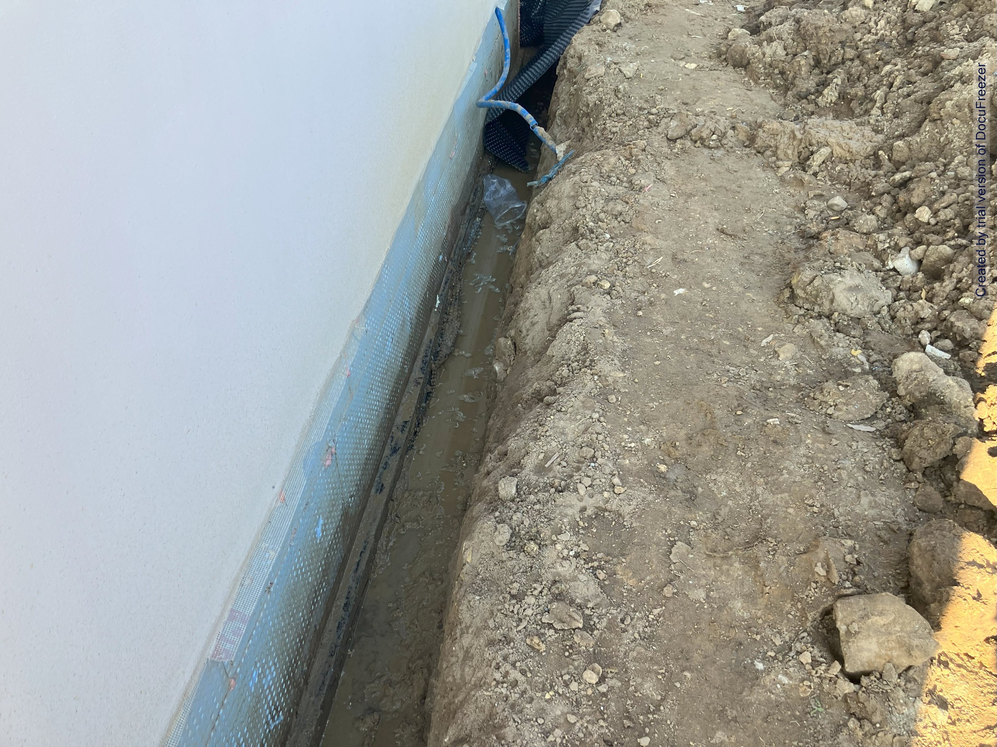 Wykopany rów wzdłuż ściany budynku z widoczną izolacją fundamentową, częściowo wypełniony wodą i z wystającymi rurami drenażowymi.