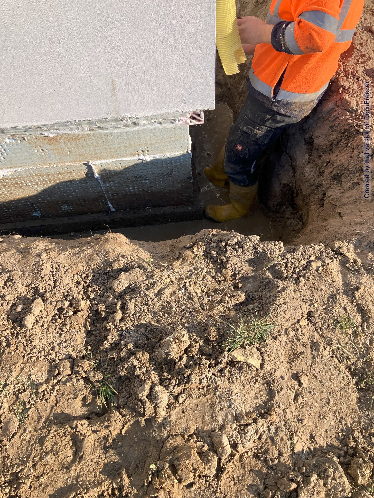 Pracownik w żółtych gumowcach stoi w wykopie przy ścianie budynku, trzymając żółty wąż; widoczna ziemia i fragment ocieplenia fundamentów.