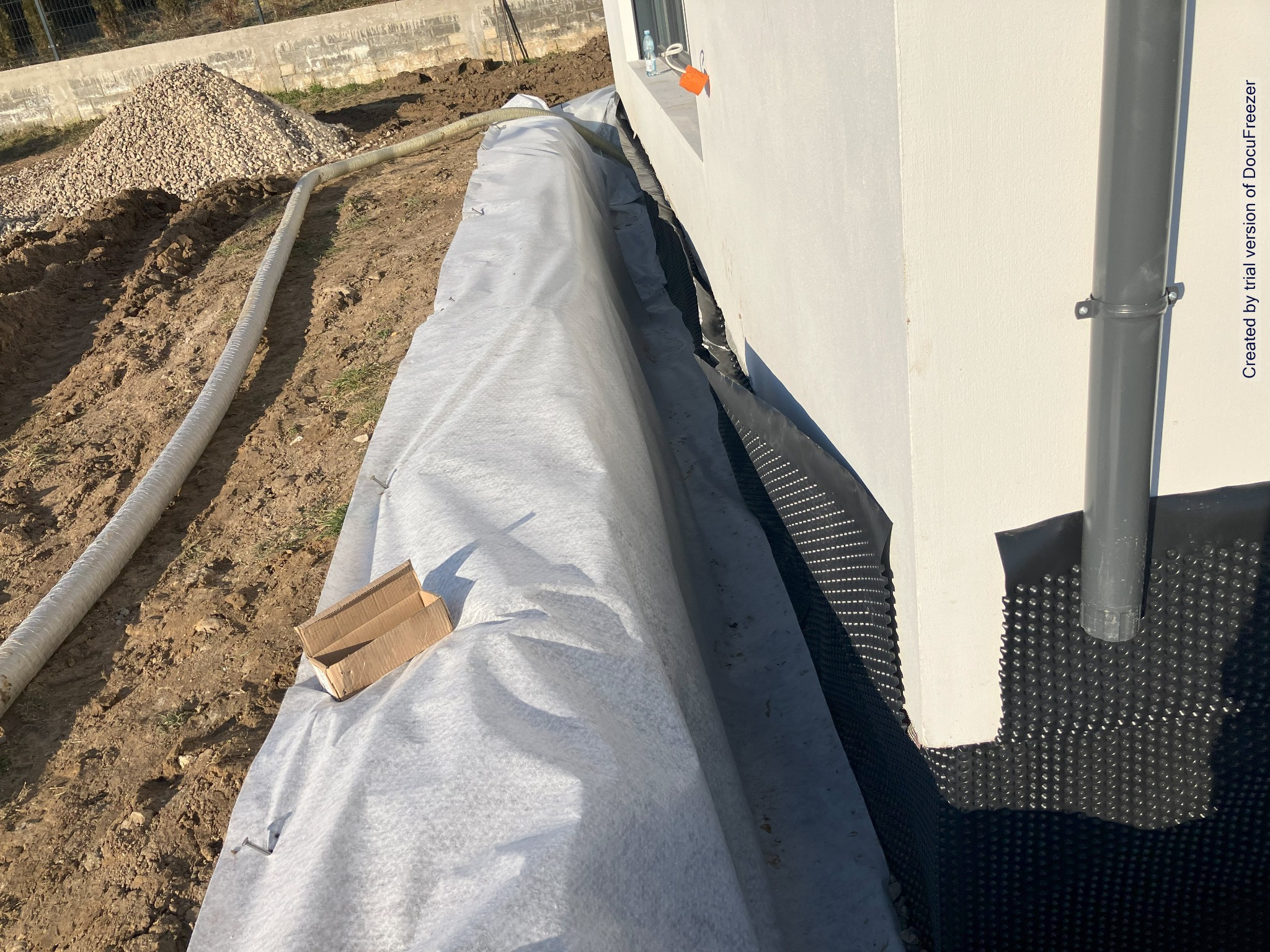Ochrona fundamentów budynku w trakcie budowy: czarna folia kubełkowa, geowłóknina, drenaż, rura drenarska i pryzma żwiru.