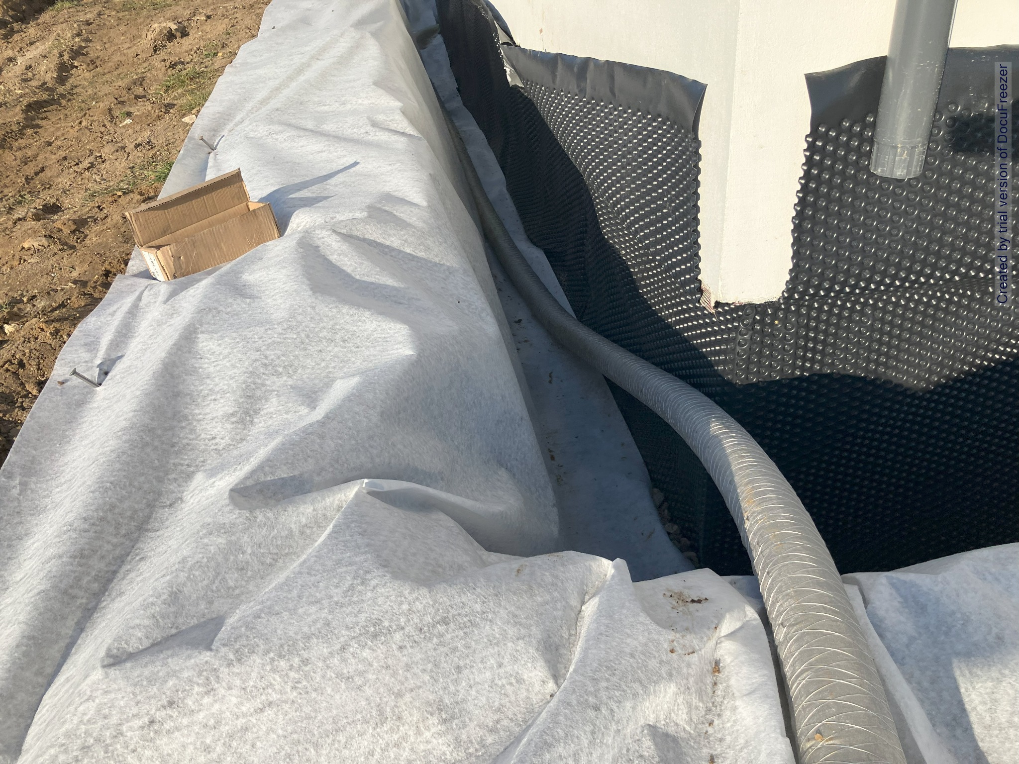 Odwodnienie fundamentów budynku w trakcie prac: biała geowłóknina zabezpiecza drenaż, widoczna rura drenarska i folia kubełkowa na ścianie fundamentowej, obok ziemia i kartonowe pudełko.