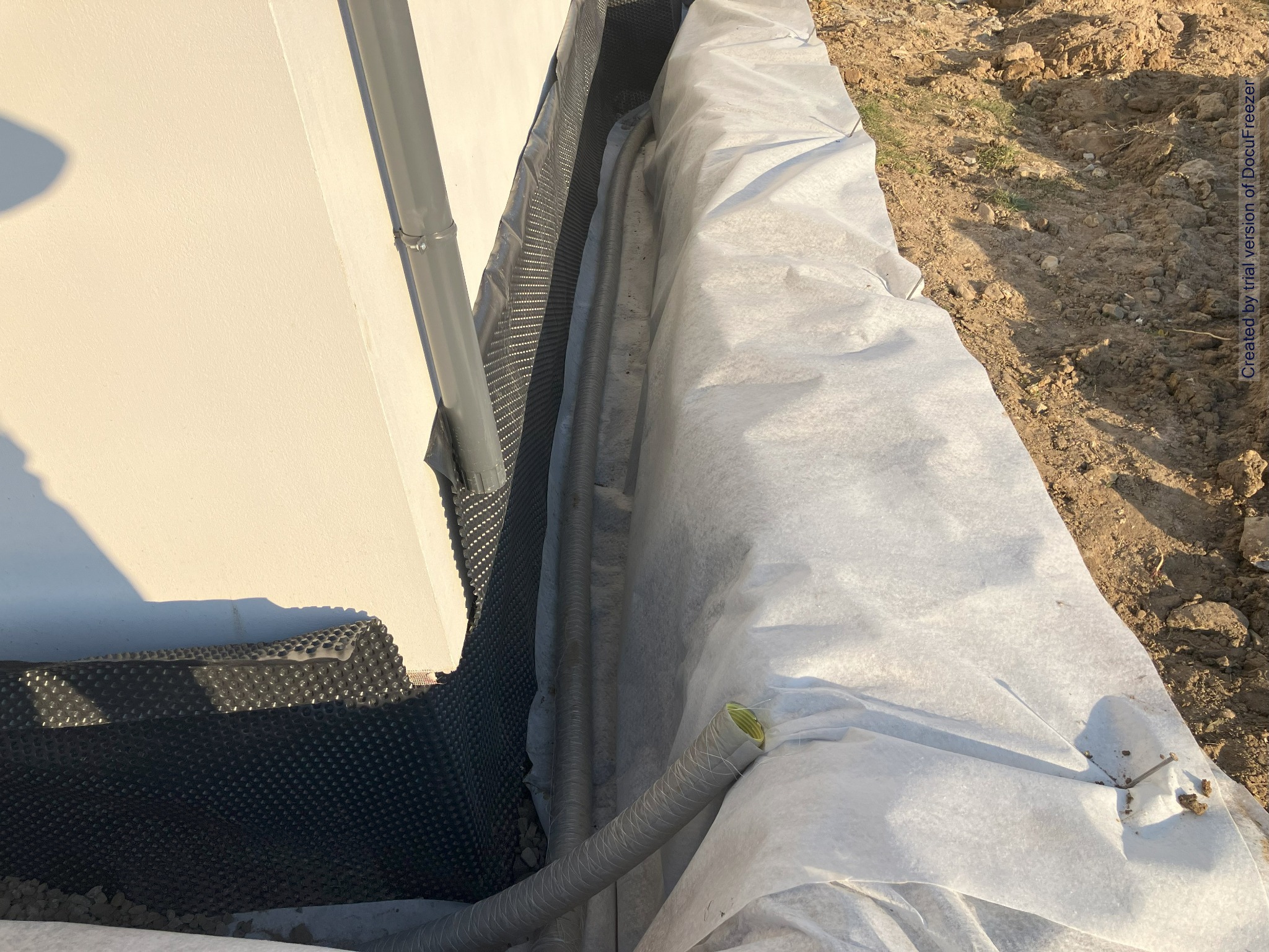 Fundament budynku w trakcie izolacji: folia kubełkowa, rury drenarskie owinięte geowłókniną, zabezpieczenie wykopu przed zasypaniem.