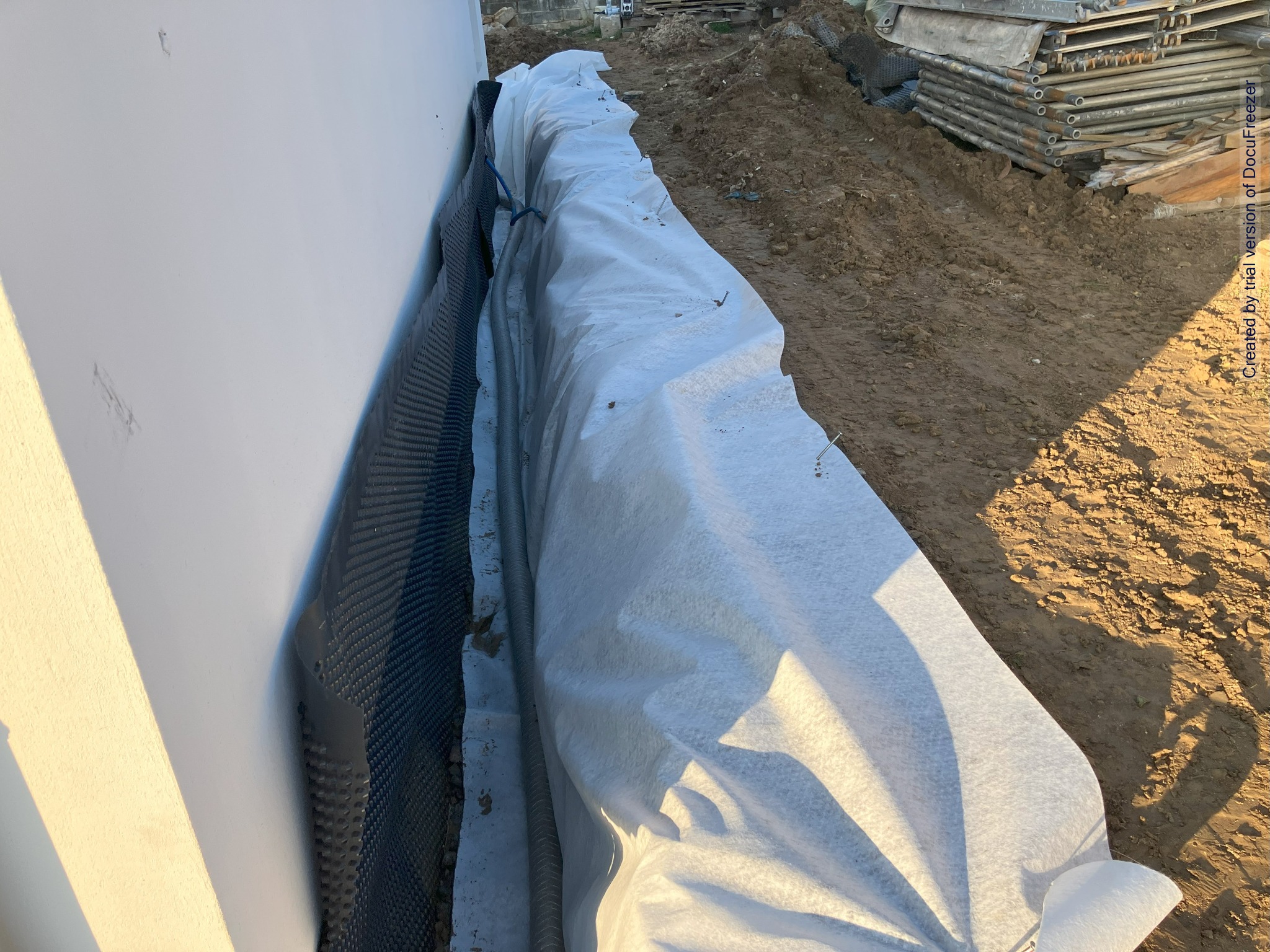 Izolacja fundamentów budynku z użyciem folii kubełkowej i geowłókniny, zabezpieczona przed zasypaniem ziemią. W tle widoczny stos rusztowań.