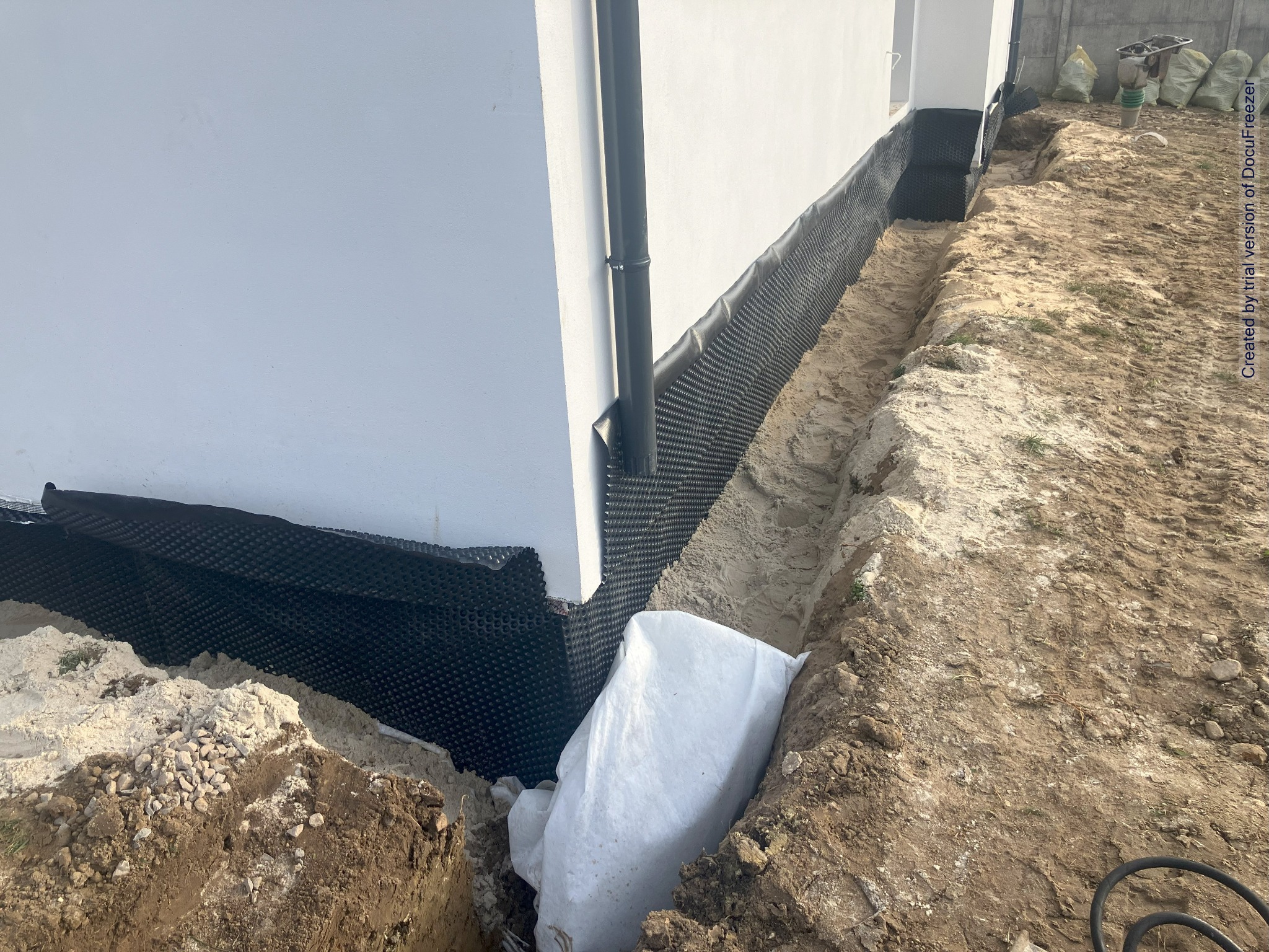Ocieplenie fundamentów budynku z użyciem folii kubełkowej, widoczny wykop, biała ściana budynku i rura spustowa, w tle zagęszczarka i worki.