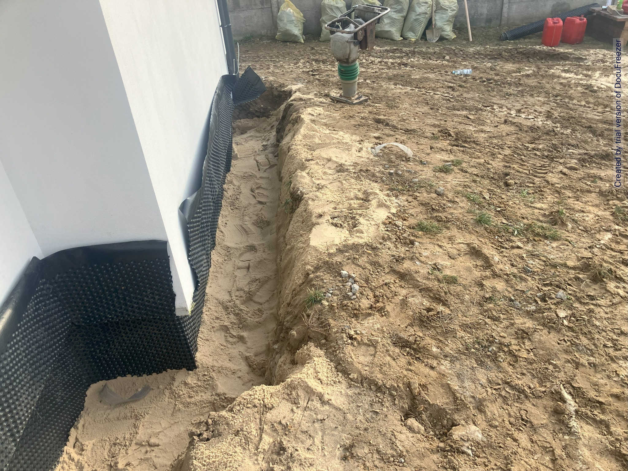 Wykop w piasku wzdłuż ściany budynku zabezpieczonej folią kubełkową, ubijarka wibracyjna na gruncie, worki z ziemią w tle.