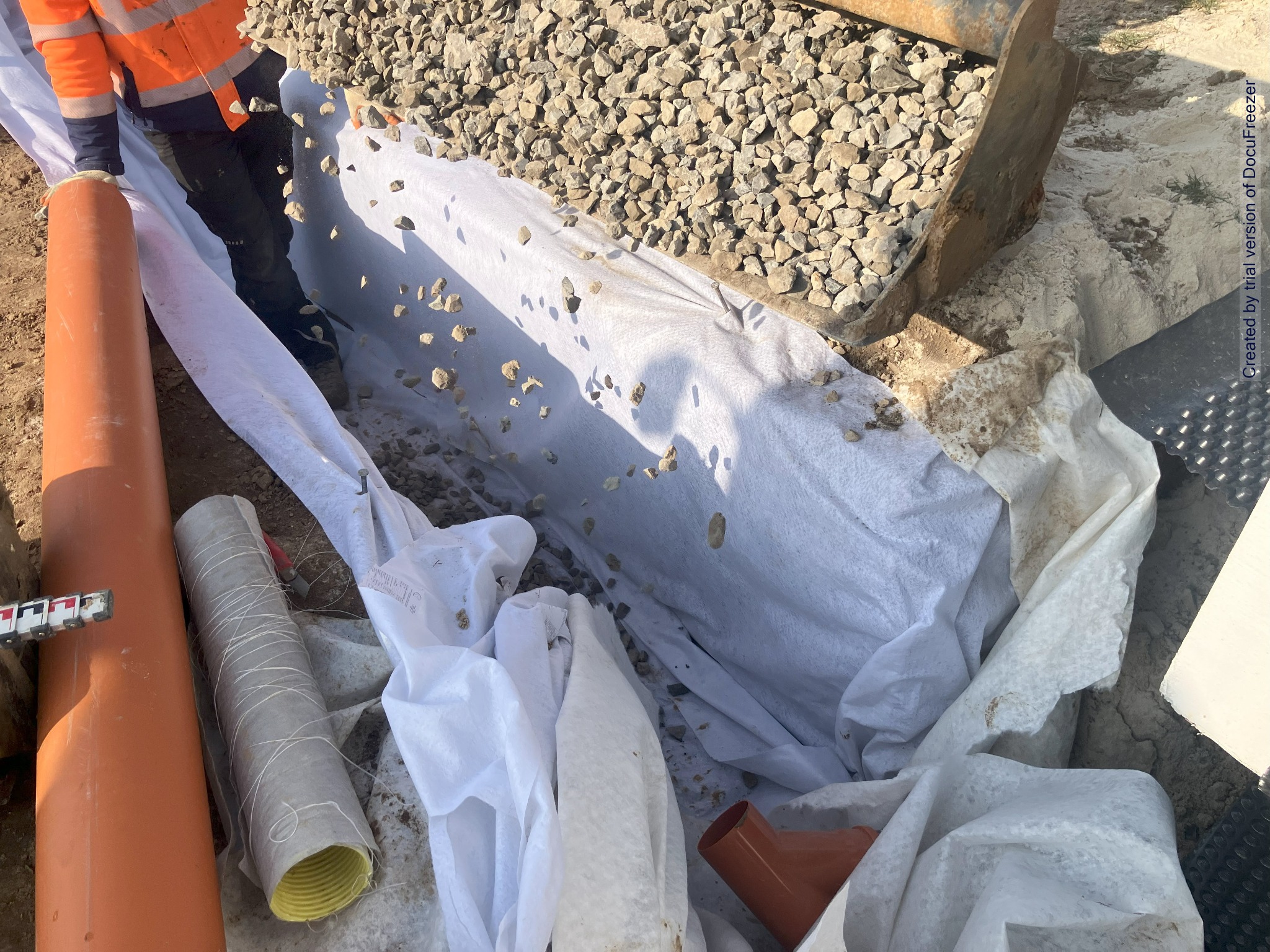 Wykop wyłożony białą geowłókniną, częściowo zasypany żwirem. Widoczne rury drenarskie, pomarańczowe złączki i fragment koparki.