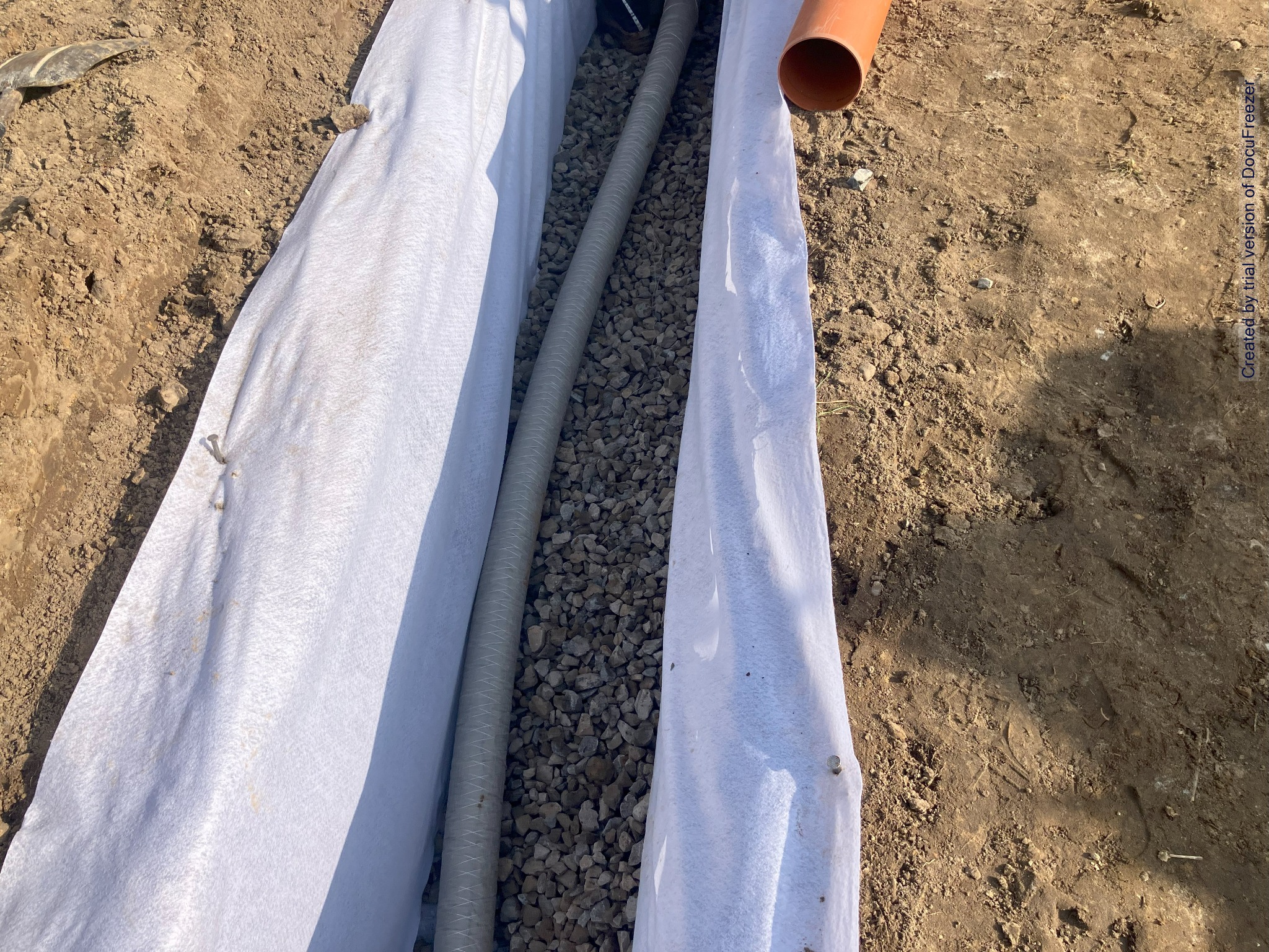 Wykop wyłożony białą geowłókniną, z rurą drenarską i warstwą drenażową z kamieni, widok z góry na etapie budowy.