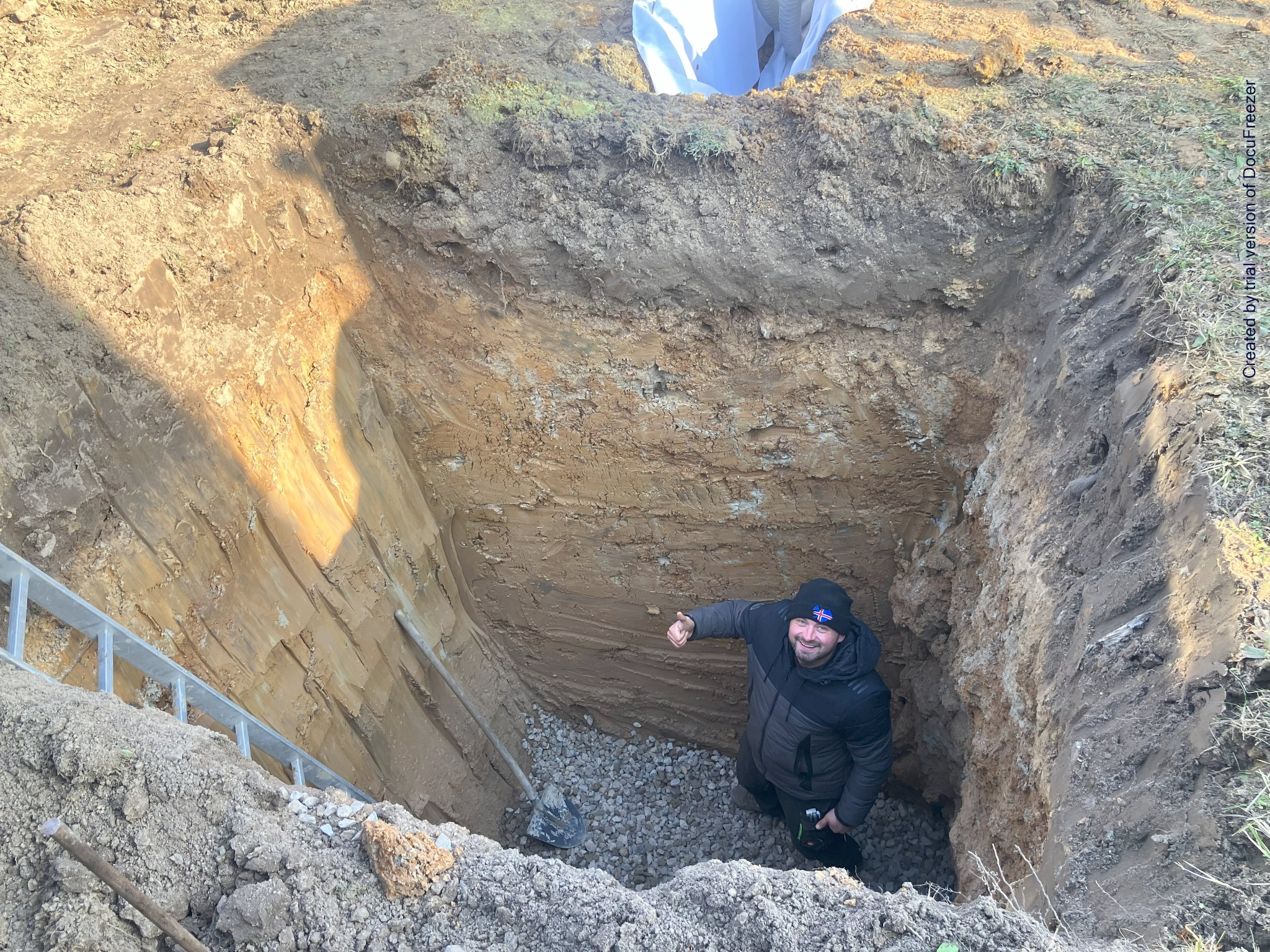 Głęboki wykop w ziemi z warstwami gleby, na dnie warstwa żwiru, mężczyzna w czapce z flagą Islandii stoi w wykopie i pokazuje kciuk w górę, drabina oparta o ścianę wykopu.