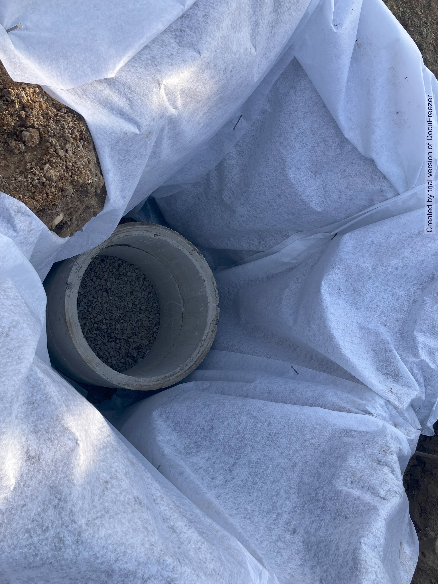 Betonowa rura wypełniona żwirem, owinięta białą włókniną, częściowo zakopana w ziemi, widoczne ślady po wbitych szpilkach mocujących włókninę.