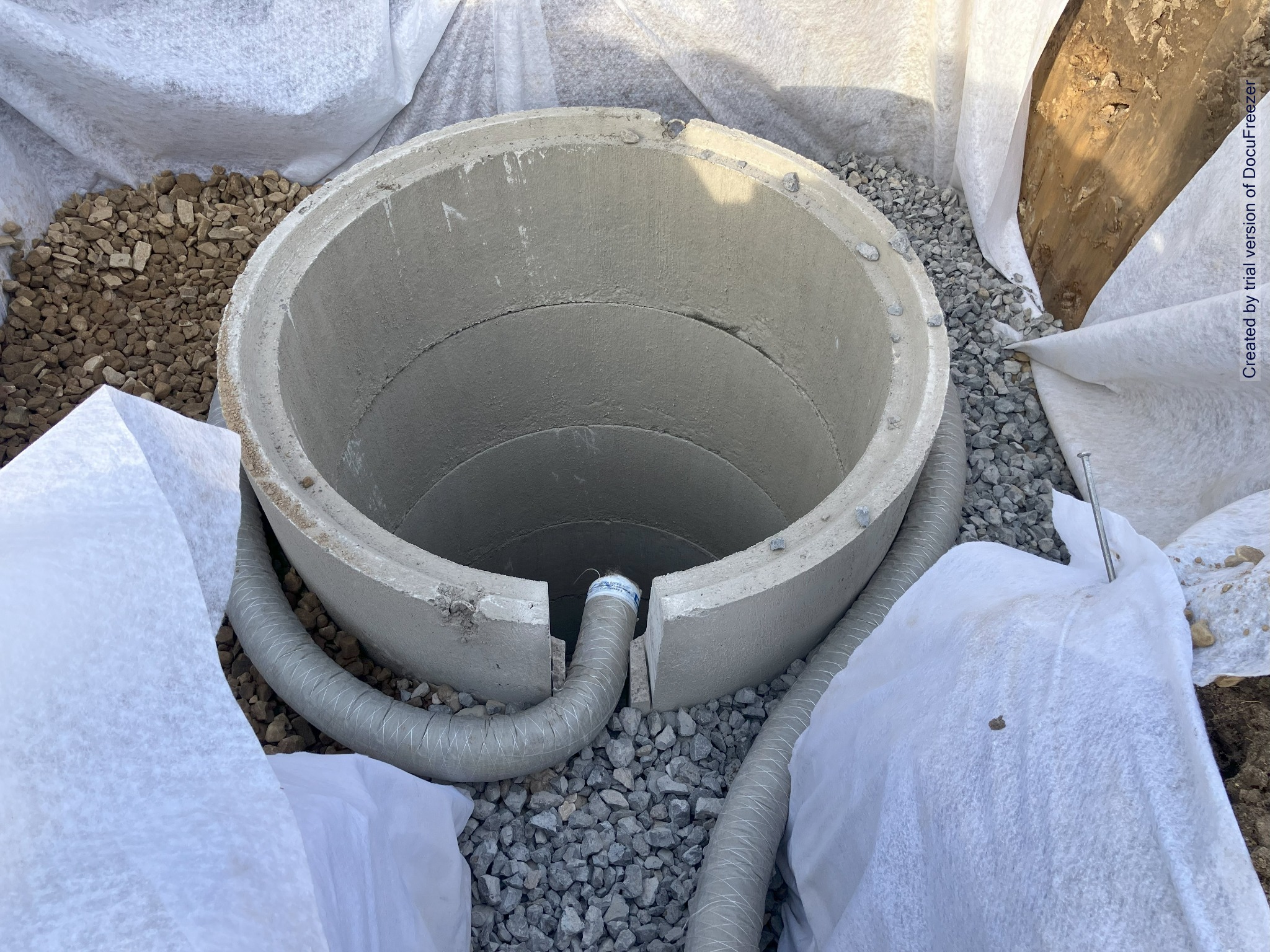 Betonowy krąg szamba w wykopie, otoczony żwirem i geowłókniną, widoczne dwa karbowane węże podłączeniowe.