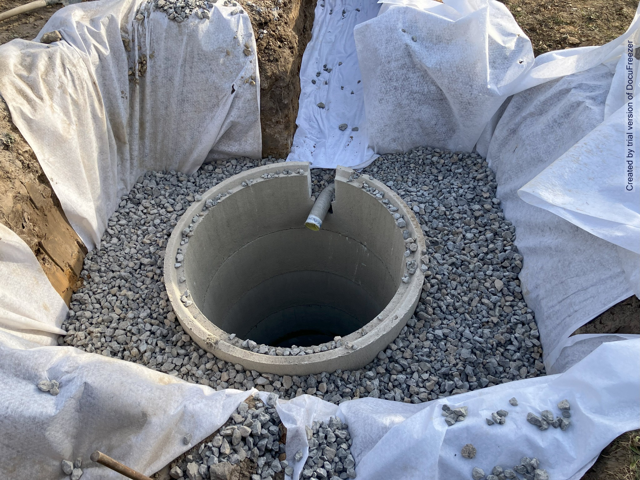 Betonowy krąg szamba w wykopie wyłożonym białą geowłókniną, otoczony warstwą żwiru, widoczny fragment rury odpływowej