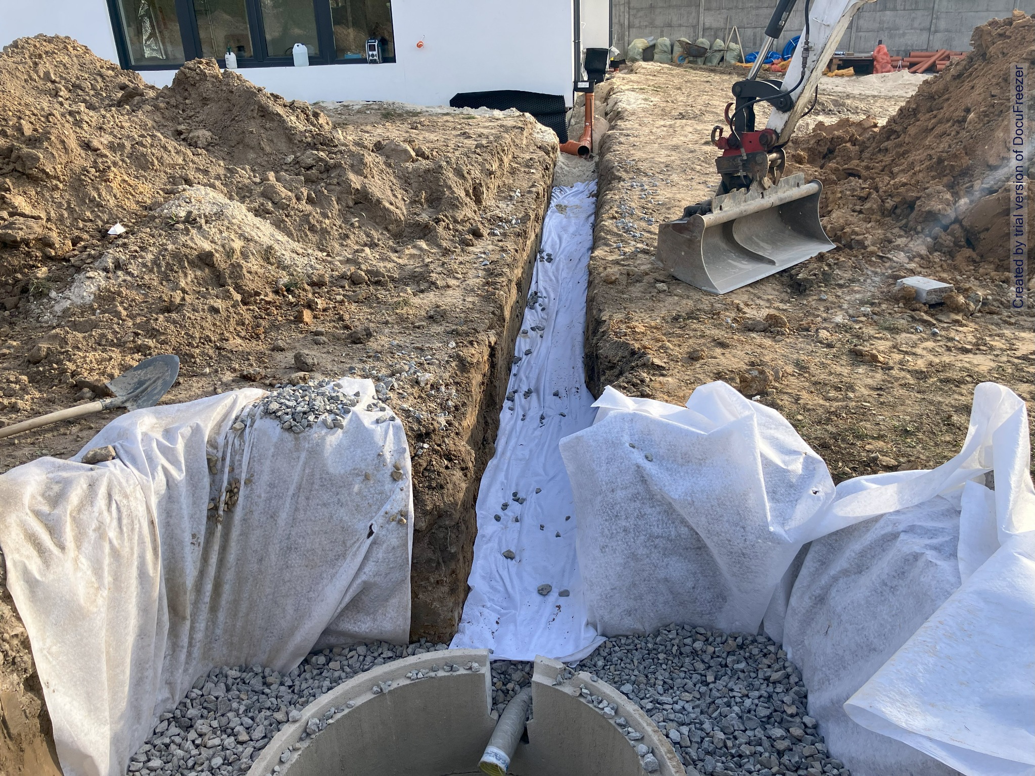 Wykop liniowy zabezpieczony geowłókniną, z widocznym drenażem obsypanym żwirem i fragmentem studzienki betonowej, obok mini koparka i pryzma ziemi.