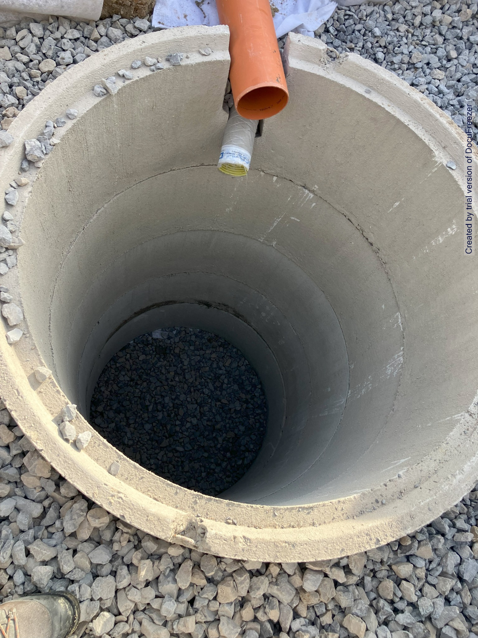 Betonowy krąg studni z wystającą rurą PCV, wypełniony gruzem, otoczony drobnym kamieniem, widok z góry.