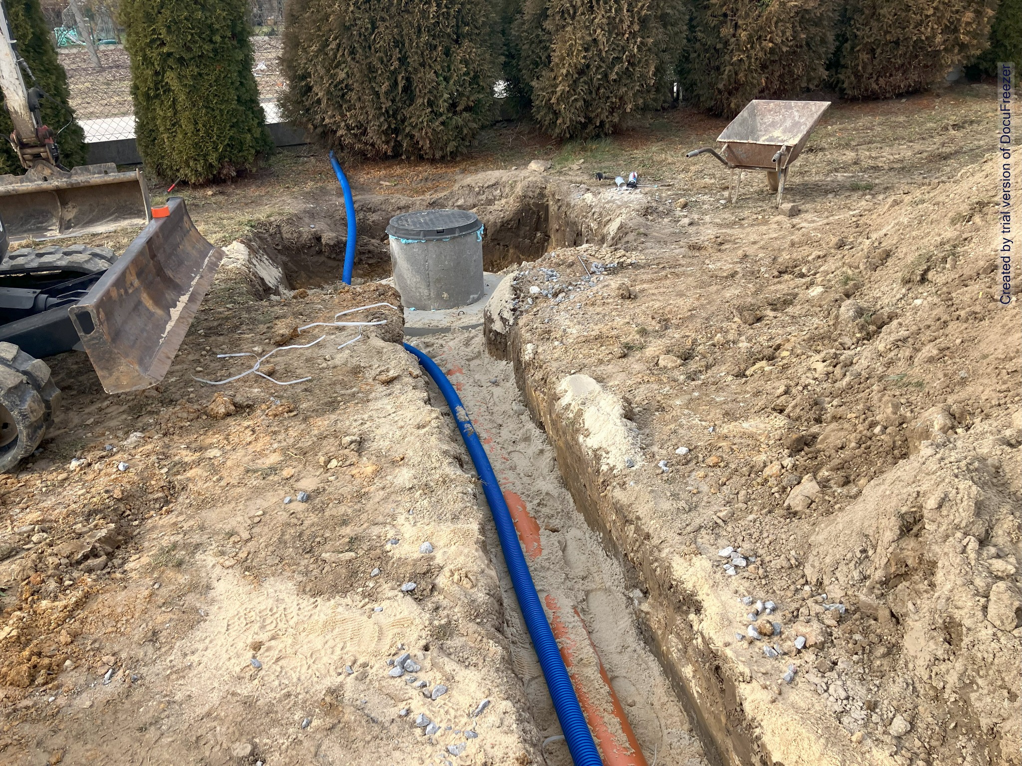 Wykop w ziemi z ułożonymi rurami instalacyjnymi w kolorze niebieskim i pomarańczowym, widoczny betonowy zbiornik z pokrywą, mini koparka i taczka w tle, teren przygotowany pod instalacje.