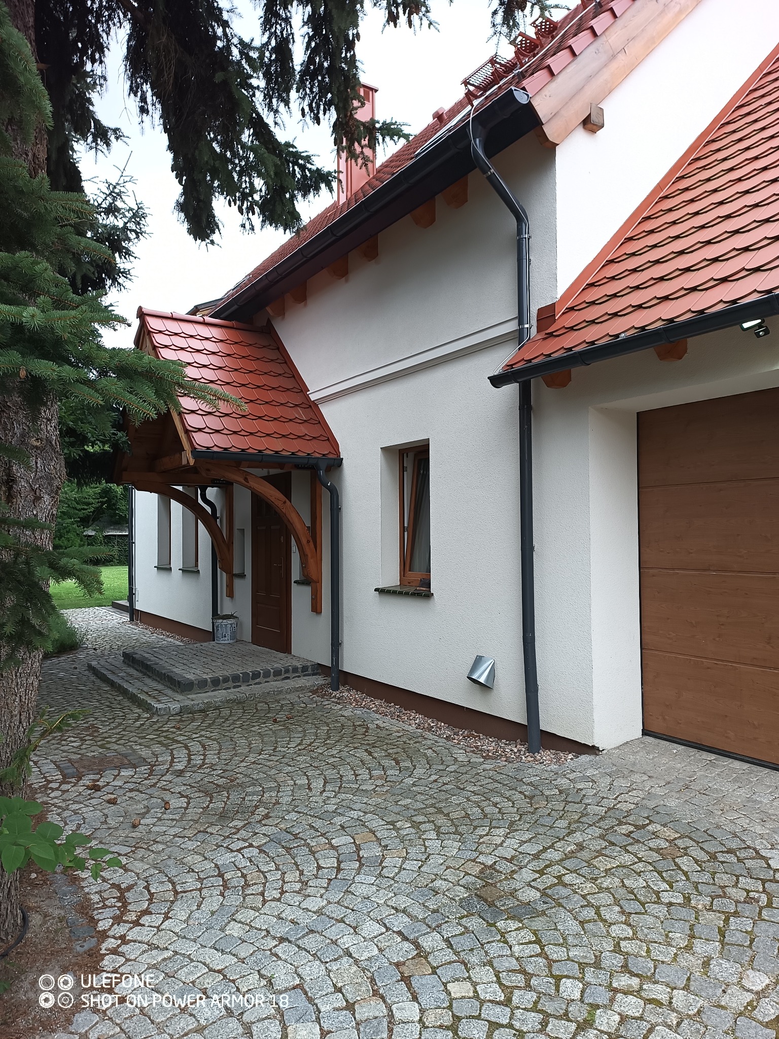 Elewacja domu jednorodzinnego z drewnianą wiatą nad wejściem, pokrytą czerwoną dachówką, widok na podjazd wyłożony kostką granitową.