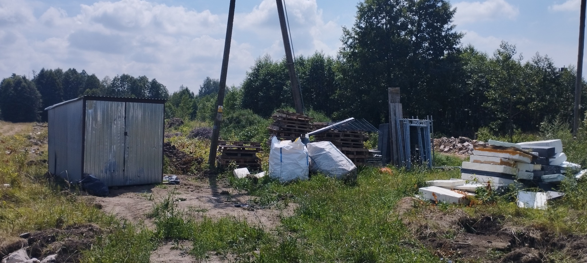 Teren budowy z metalowym garażem, paletami, workami i stertą styropianu na tle drzew i nieba.