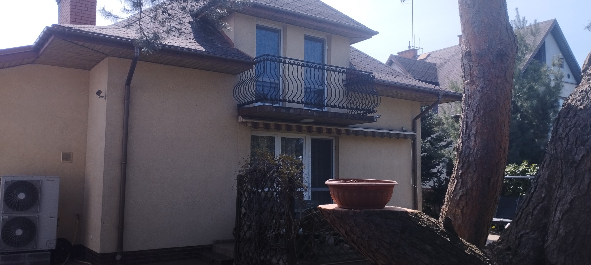 Widok z zewnątrz na dom z beżową elewacją, balkonem z ozdobną, czarną balustradą i klimatyzacją na ścianie, z pniem drzewa w pierwszym planie, na którym stoi donica.