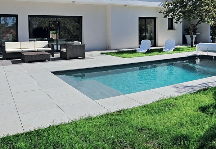 Nowoczesny, prostokątny basen z płytkimi schodami wejściowymi, otoczony jasnoszarą płytką i trawnikiem, z białym domem i meblami ogrodowymi w tle.