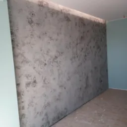 Dekoracyjna ściana w odcieniach szarości z efektem strukturalnym, świeżo po wykonaniu, w jasnym pomieszczeniu. Widoczny fragment podłogi i ściany bocznej w kolorze miętowym.