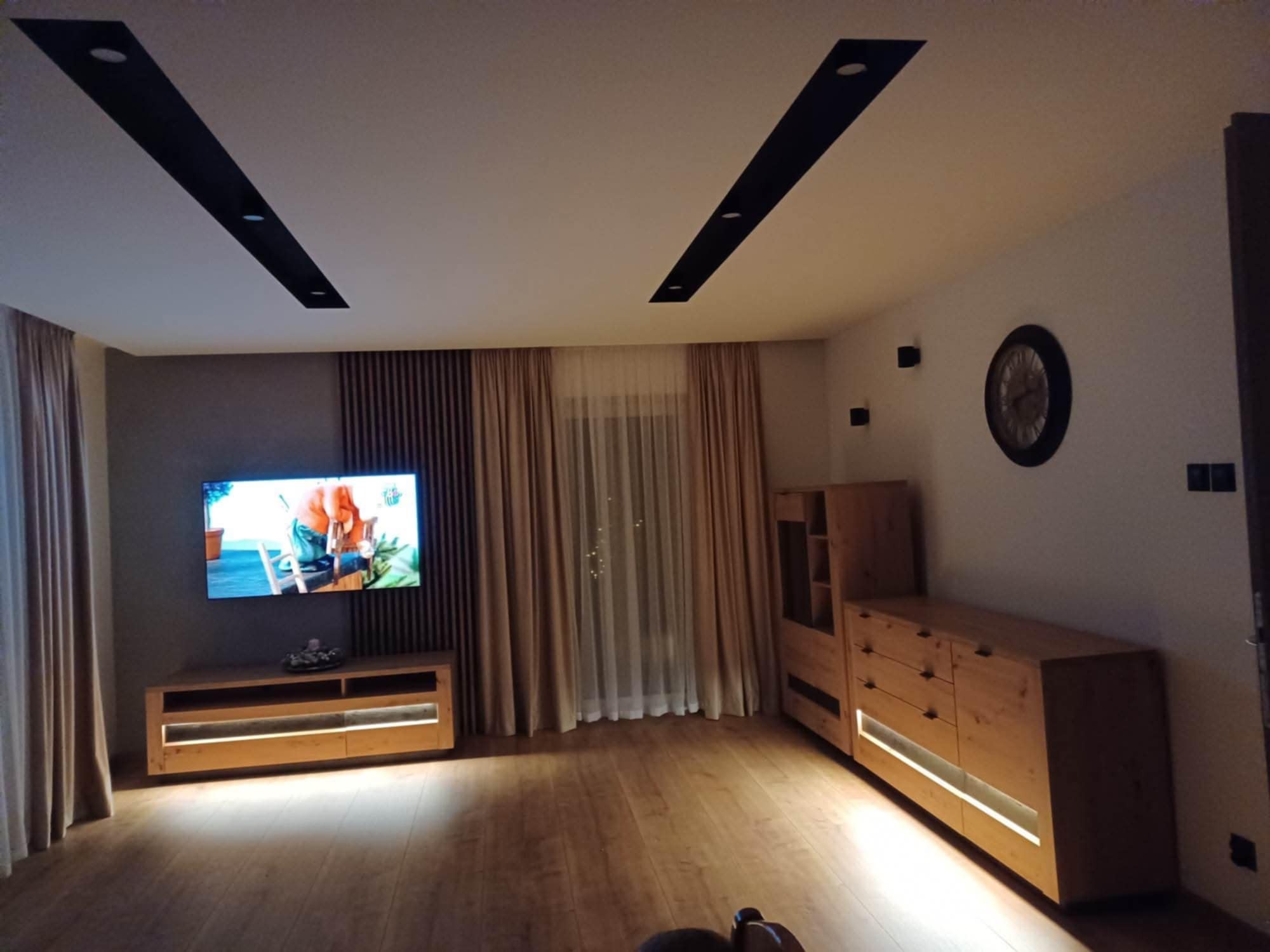 Salon z drewnianymi meblami podświetlonymi LED, ciemnymi listwami sufitowymi i zasłonami w odcieniach beżu. Na ścianie telewizor i zegar.
