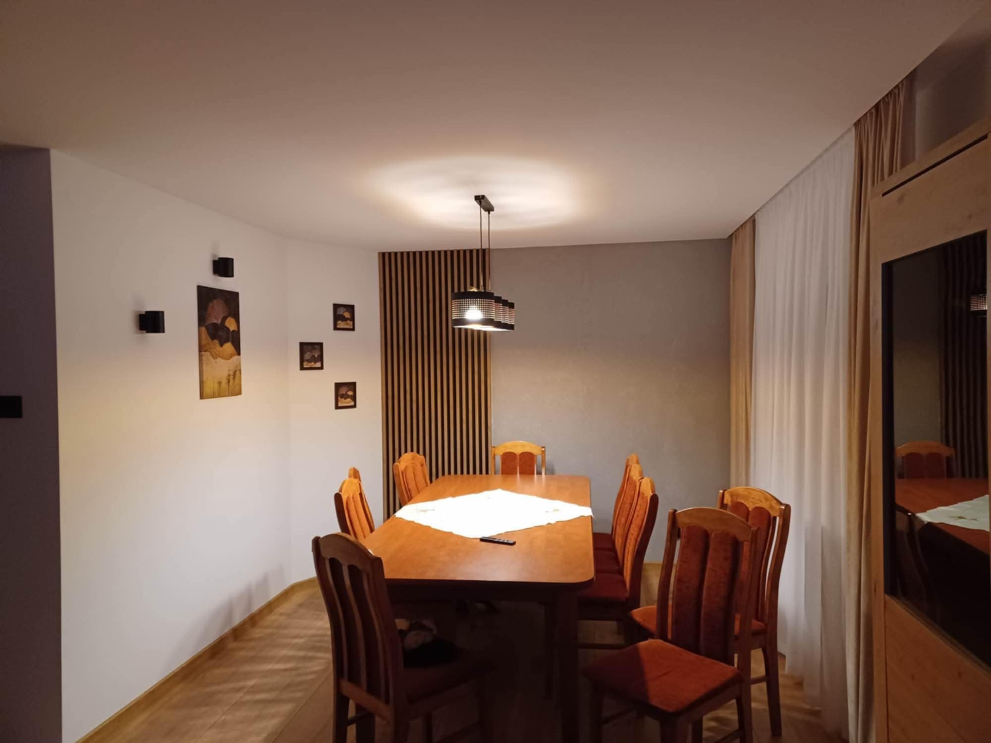 Jasna jadalnia z drewnianym stołem i krzesłami. Na ścianie dekoracyjne panele pionowe i obrazy. Centralnie umieszczona lampa sufitowa. Widoczne drewniane wykończenia.