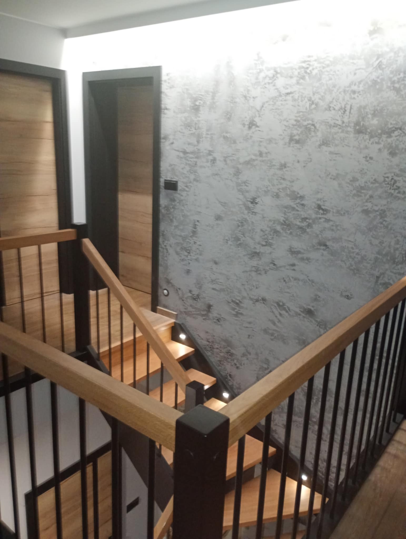 Schody drewniane z czarnymi balustradami i oświetleniem punktowym, ściana z dekoracyjną strukturą w odcieniach szarości. Widok z góry, drzwi z okleiną drewnopodobną w tle.