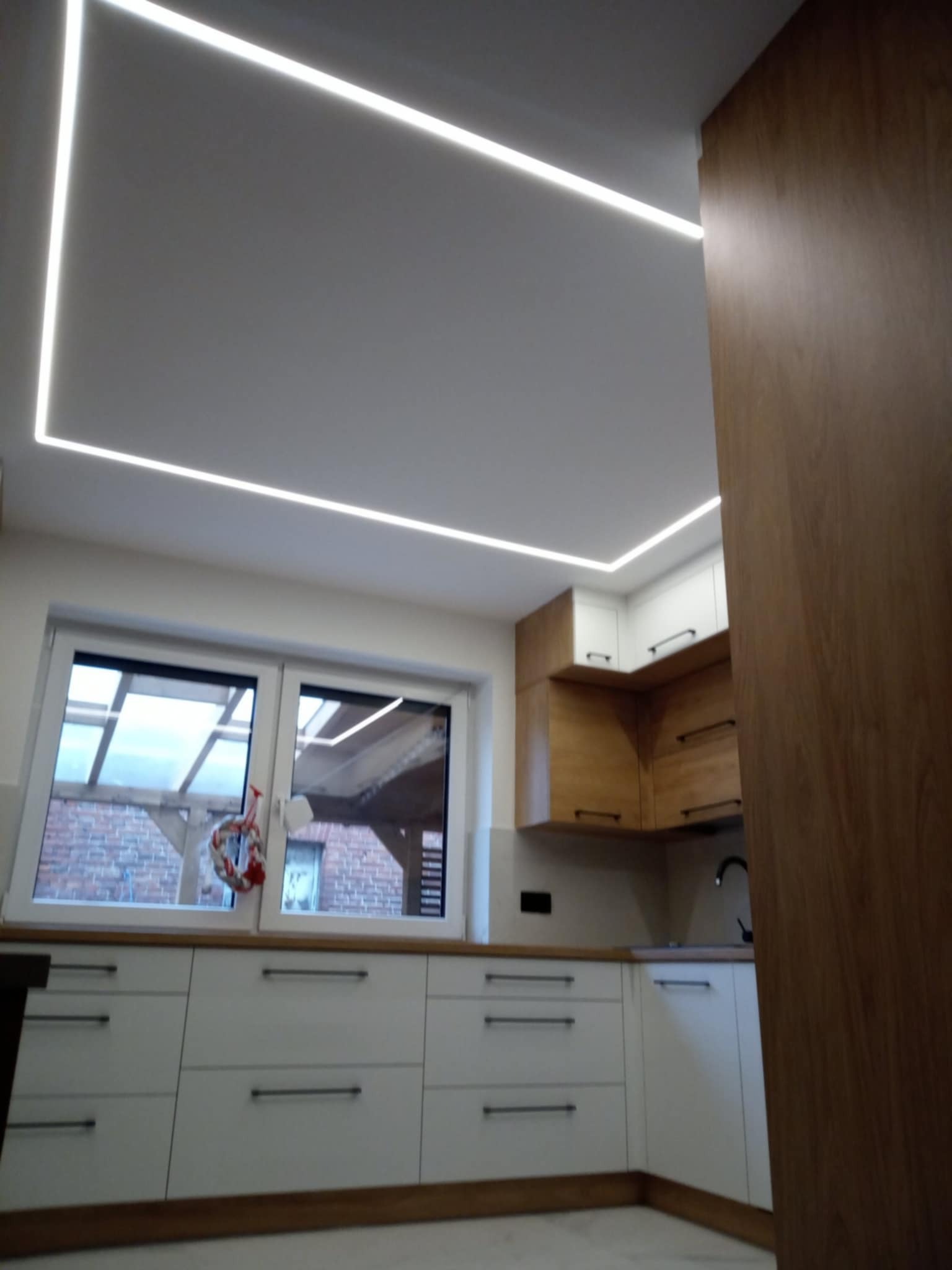 Nowoczesna kuchnia z białymi szafkami i oświetleniem LED w kształcie prostokąta na suficie. Widok na okno z dekoracją świąteczną, drewniane blaty i szafki wiszące.