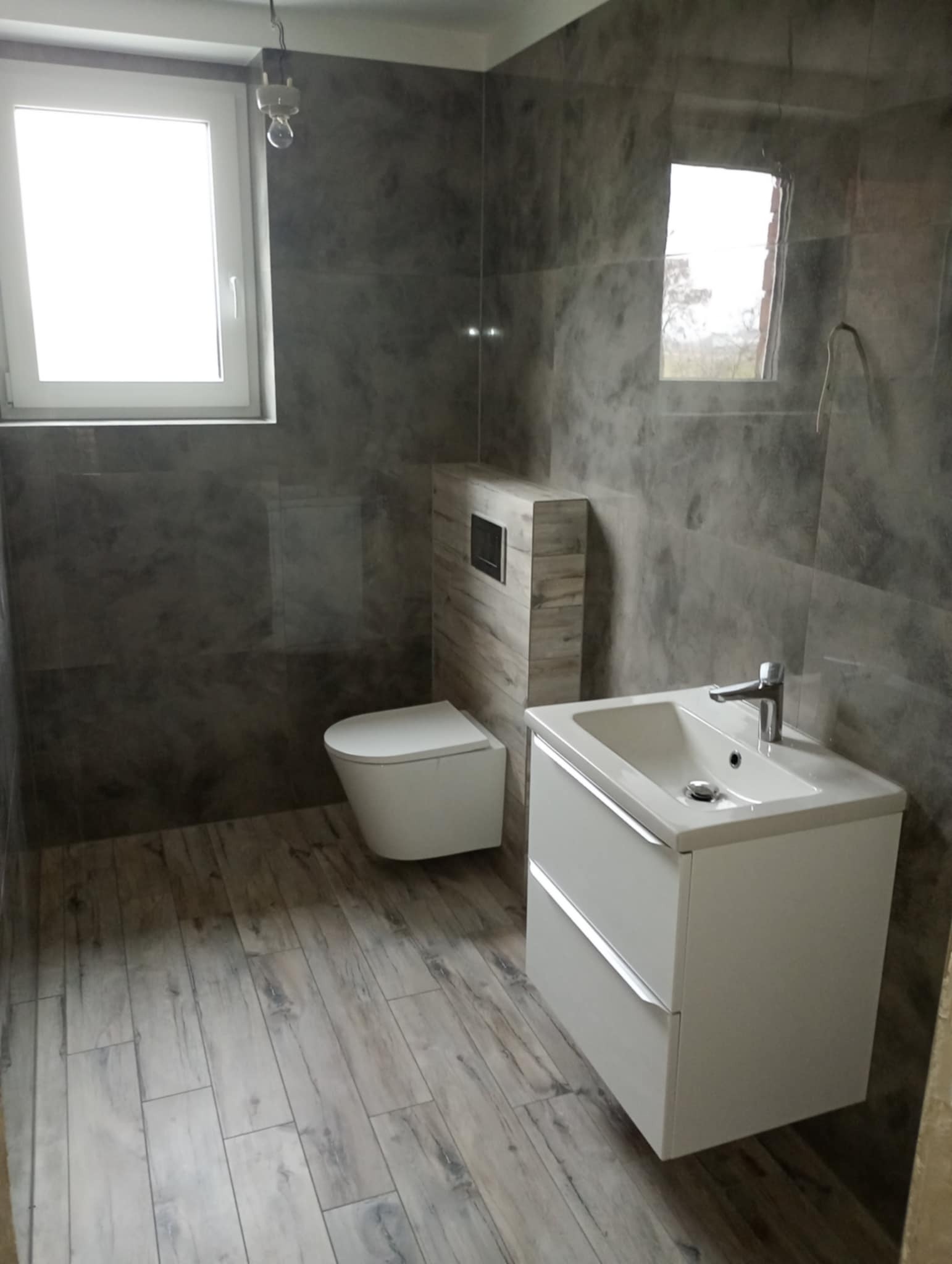 Wykończona łazienka z szarymi płytkami imitującymi marmur na ścianach i drewnopodobnymi na podłodze. Biała umywalka z szafką i toaleta.