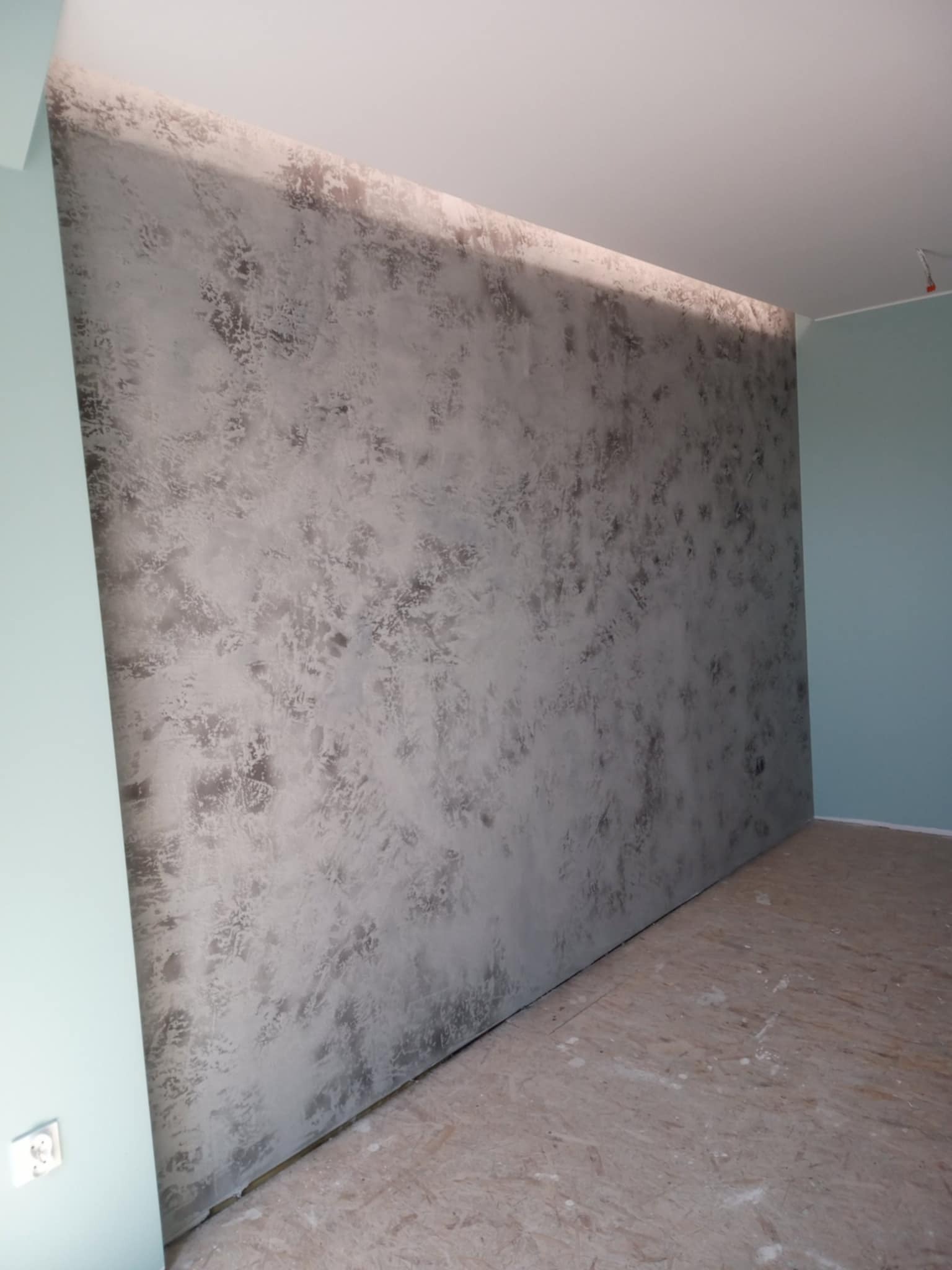 Dekoracyjna ściana w odcieniach szarości z efektem strukturalnym, świeżo po wykonaniu, w jasnym pomieszczeniu. Widoczny fragment podłogi i ściany bocznej w kolorze miętowym.