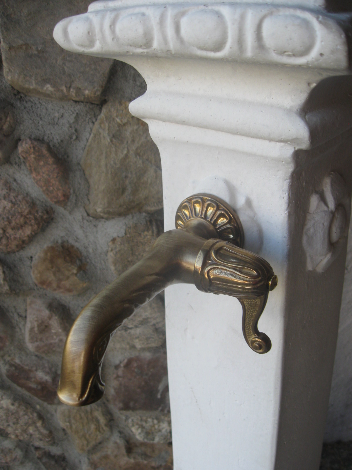 Mosiężny kran ogrodowy w stylu retro zamontowany na białym, ozdobnym postumencie z kamiennym tłem.