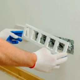 Elektryk w białych rękawicach z niebieskimi palcami montuje ramkę z czterema otworami na gniazdka elektryczne w ścianie.